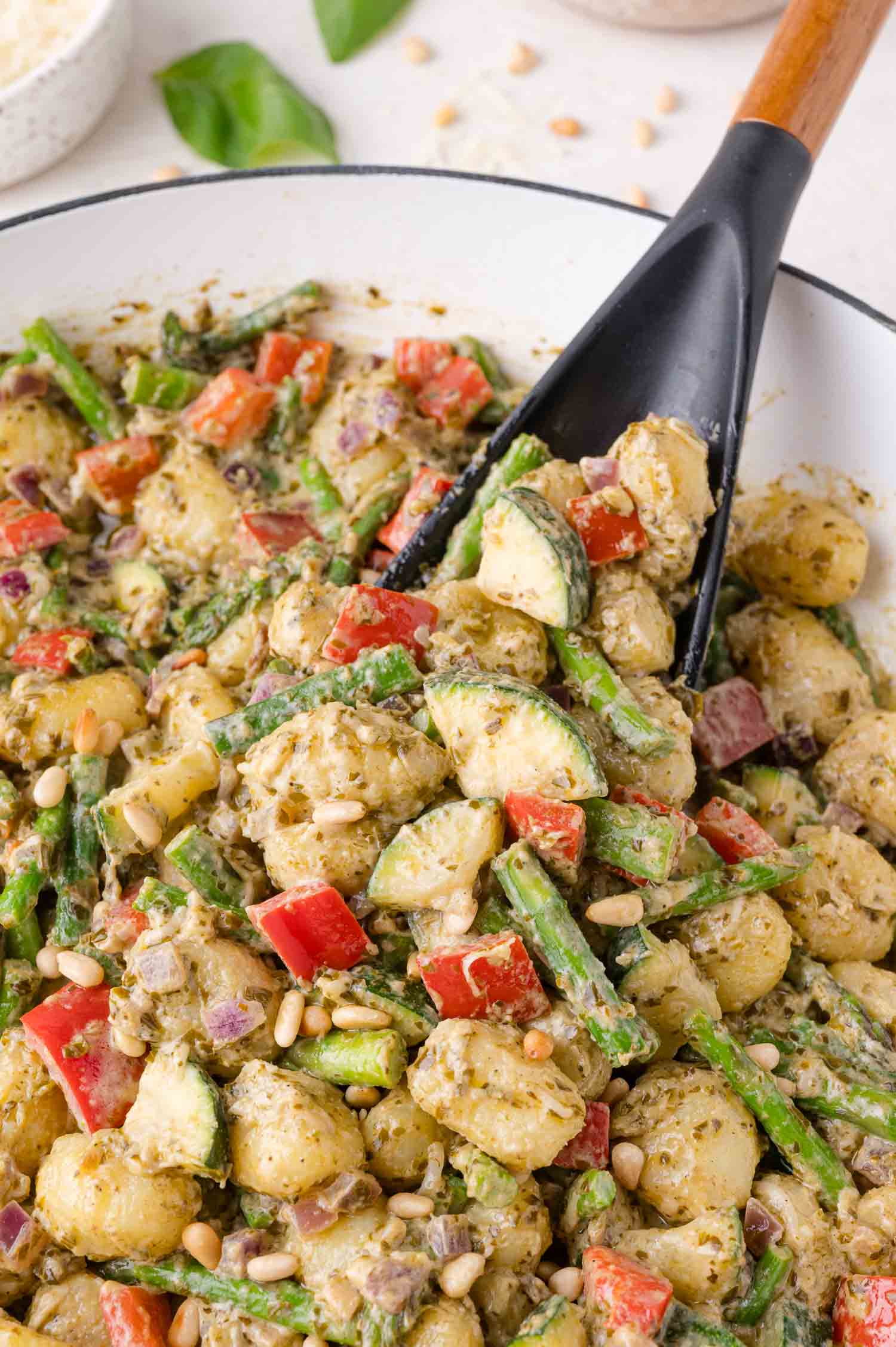 A bowl of gnocchi mixed with chopped red peppers, zucchini, asparagus, red onion, pine nuts, and coated in a green pesto sauce, with a serving spoon.