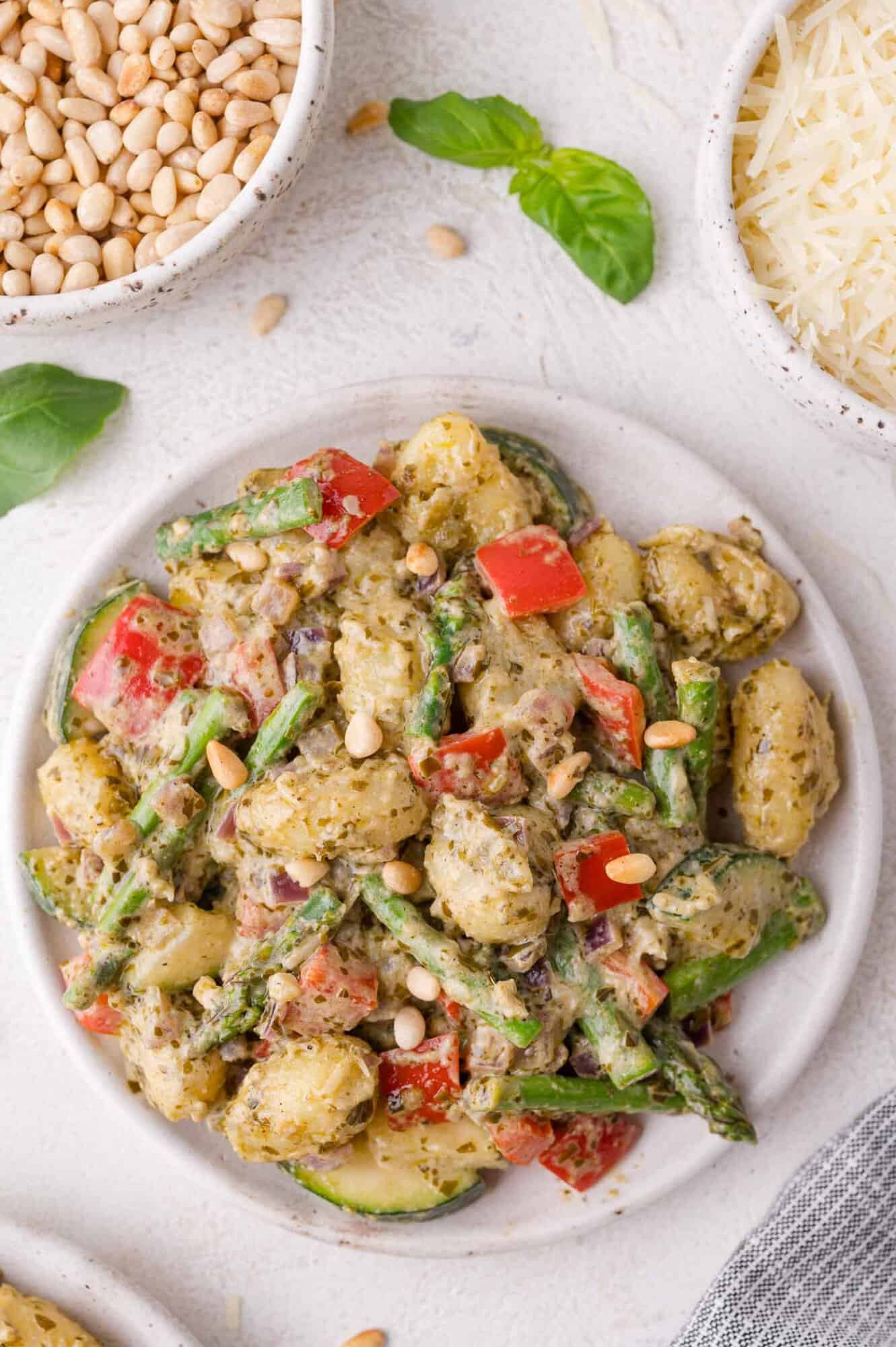A plate of gnocchi mixed with pesto, asparagus, red bell peppers, zucchini, and topped with pine nuts, with bowls of pine nuts and grated cheese nearby.