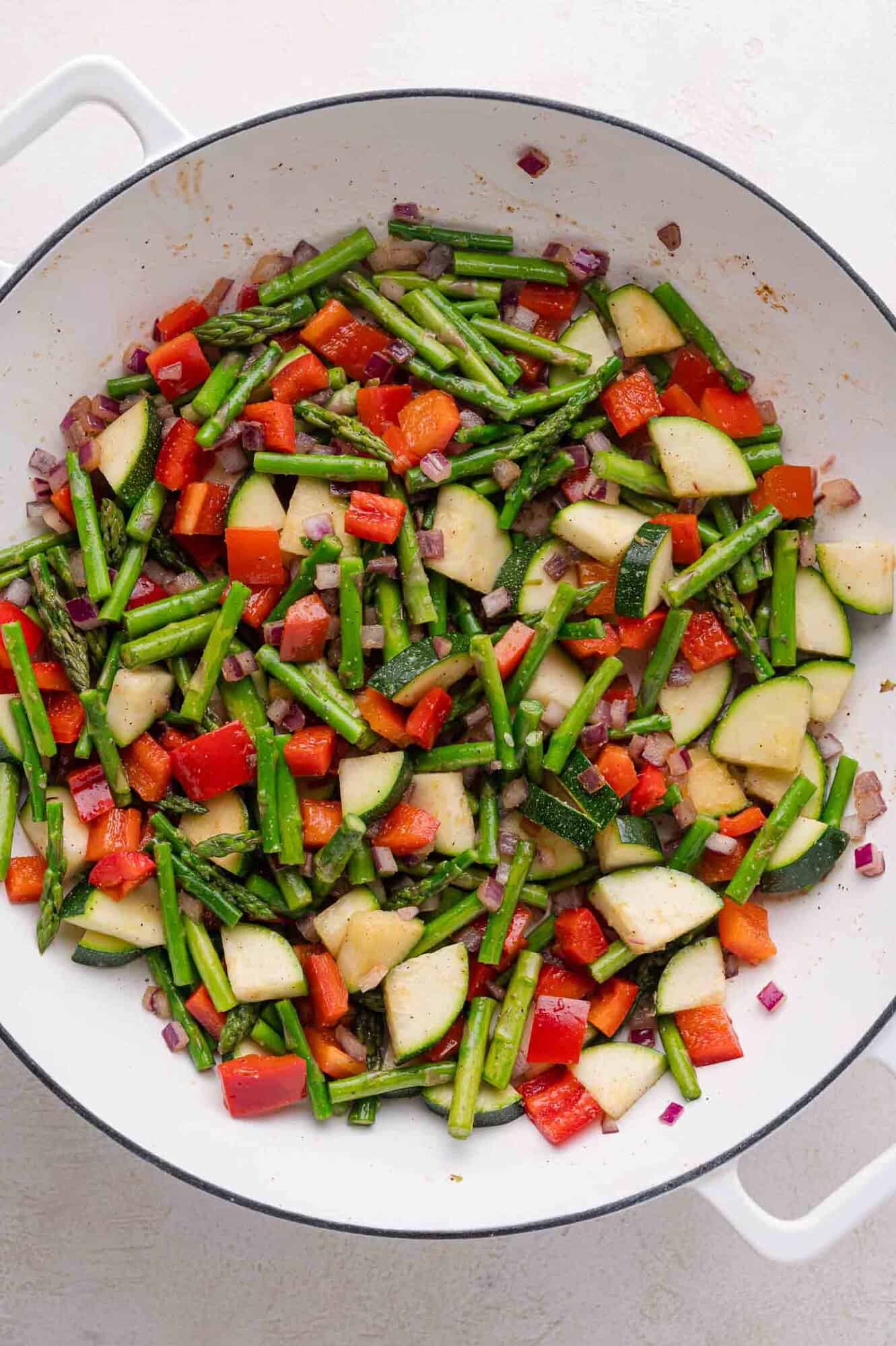A white pan filled with sautéed vegetables including asparagus, red bell pepper, zucchini, and red onion.