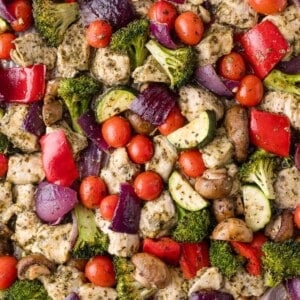 Sheet pan with cooked chicken pieces, broccoli, cherry tomatoes, red bell pepper, red onion, zucchini, and mushrooms, all seasoned with pesto and mixed together.
