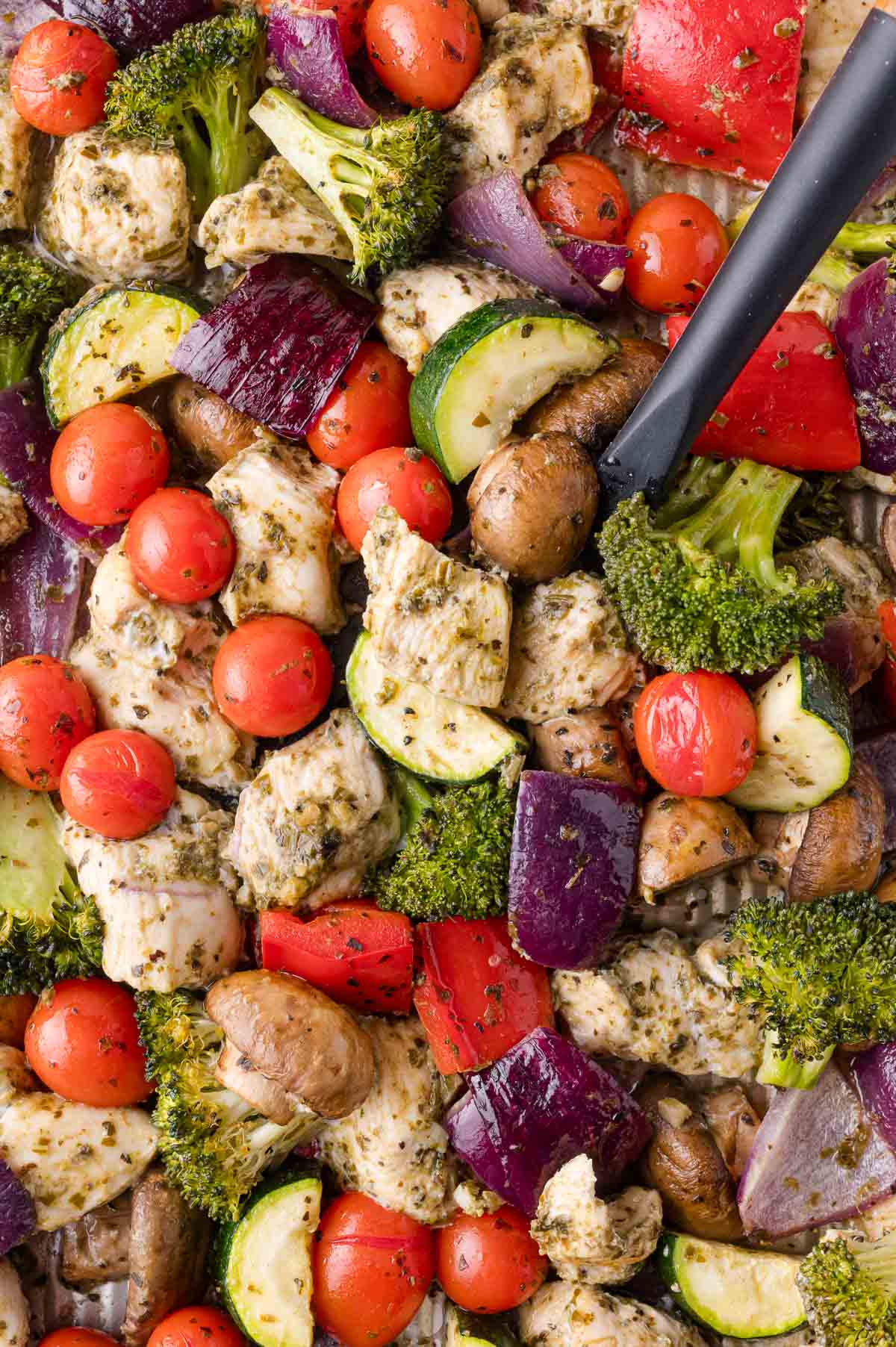 Sheet pan with baked chicken cubes and vegetables, all seasoned with pesto and roasted. A black spatula is visible on the right side.
