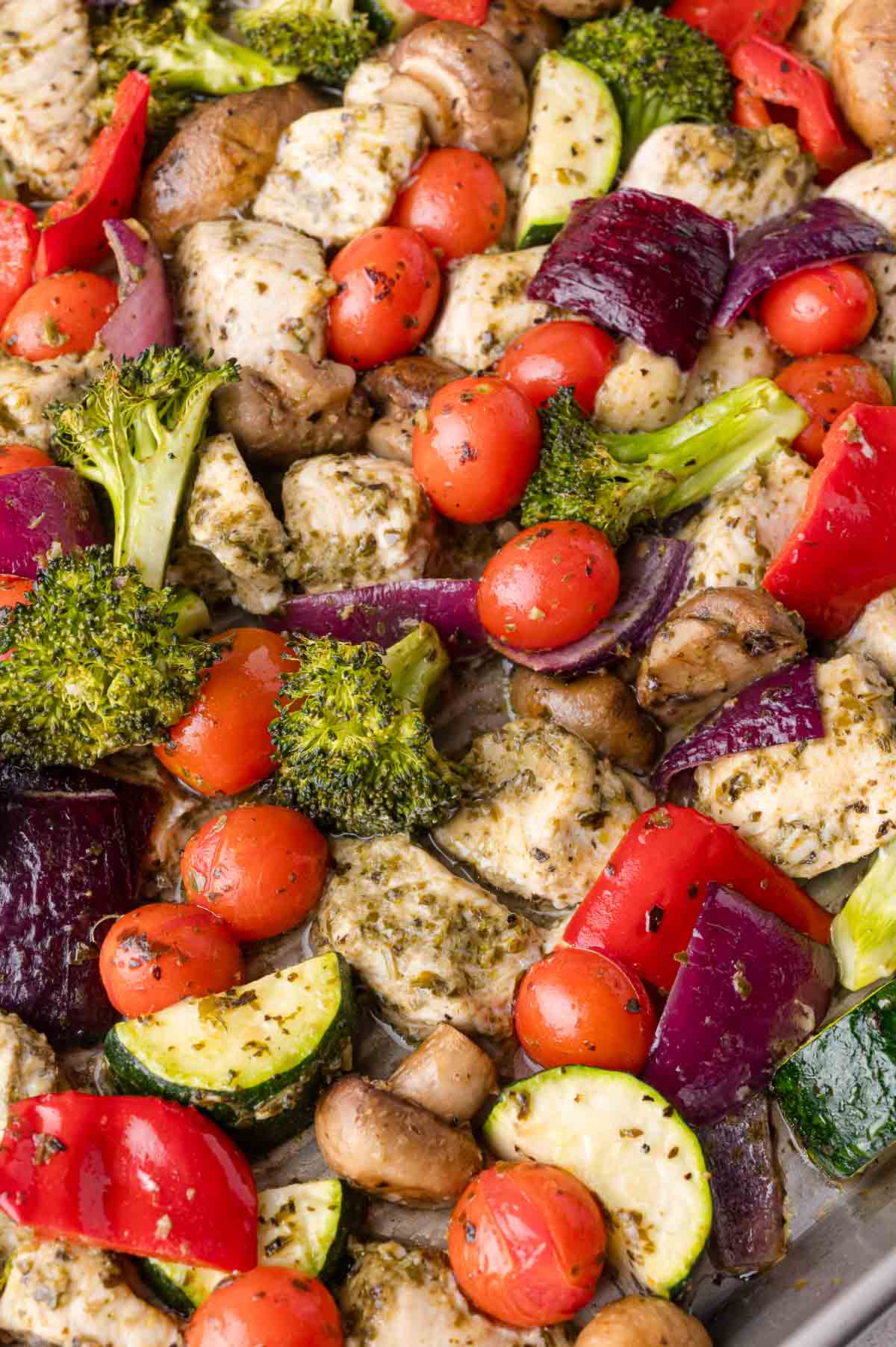 A sheet pan filled with cooked chicken pieces, cherry tomatoes, broccoli, zucchini, mushrooms, red onion, and red bell peppers, all seasoned with herbs.
