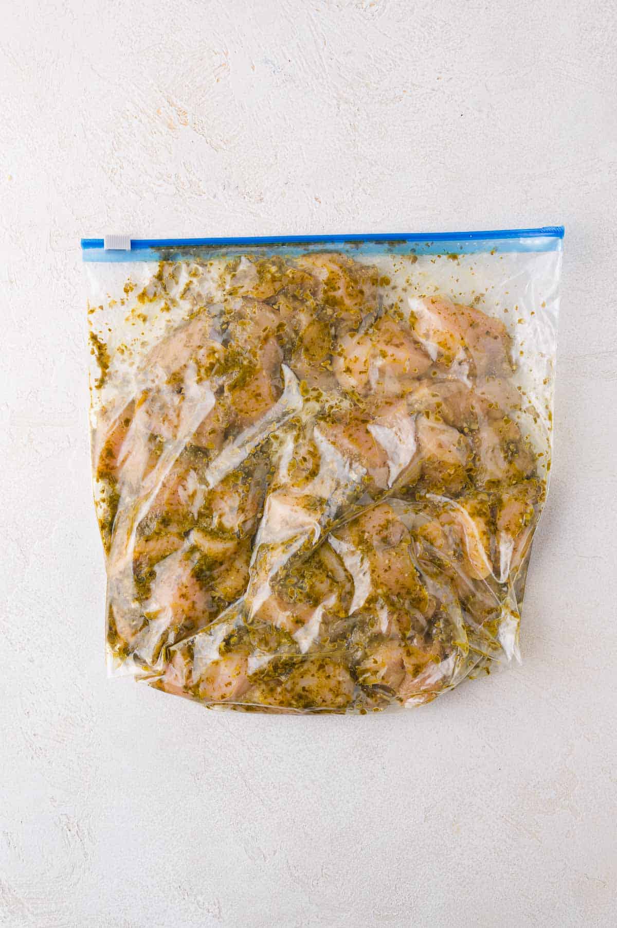 Raw chicken pieces marinating in a clear resealable plastic bag with herbs and spices, placed on a light-colored surface.