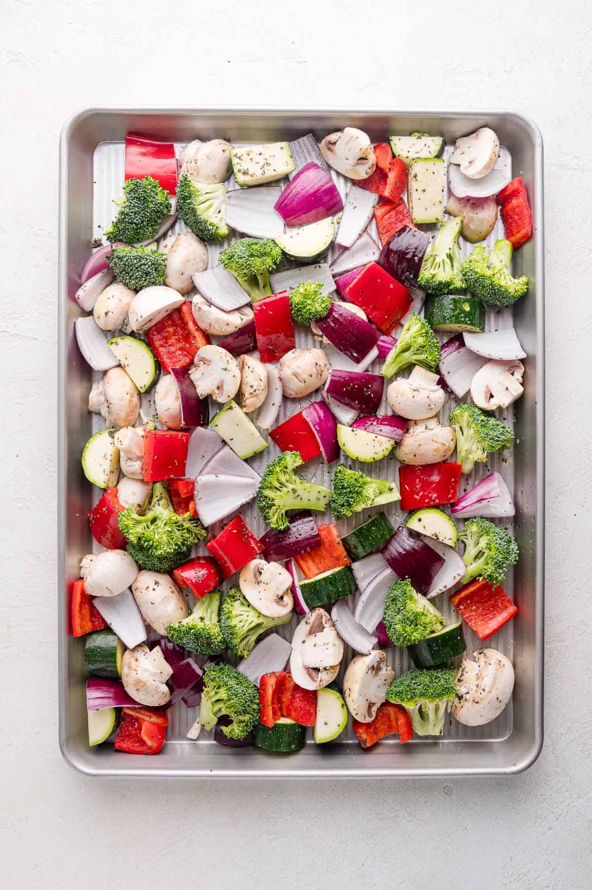 A baking sheet filled with chopped vegetables including mushrooms, broccoli, red onion, zucchini, and red bell pepper, seasoned with herbs and pepper.