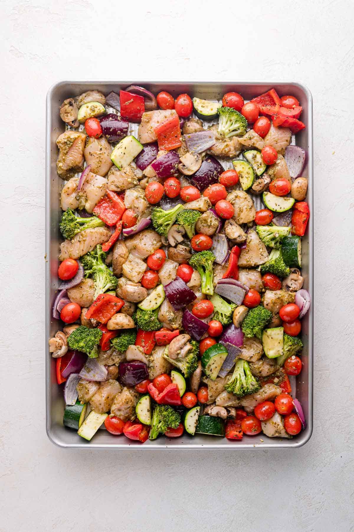 A baking tray filled with chopped chicken, broccoli, zucchini, red bell peppers, cherry tomatoes, and red onions, all seasoned and ready for roasting.
