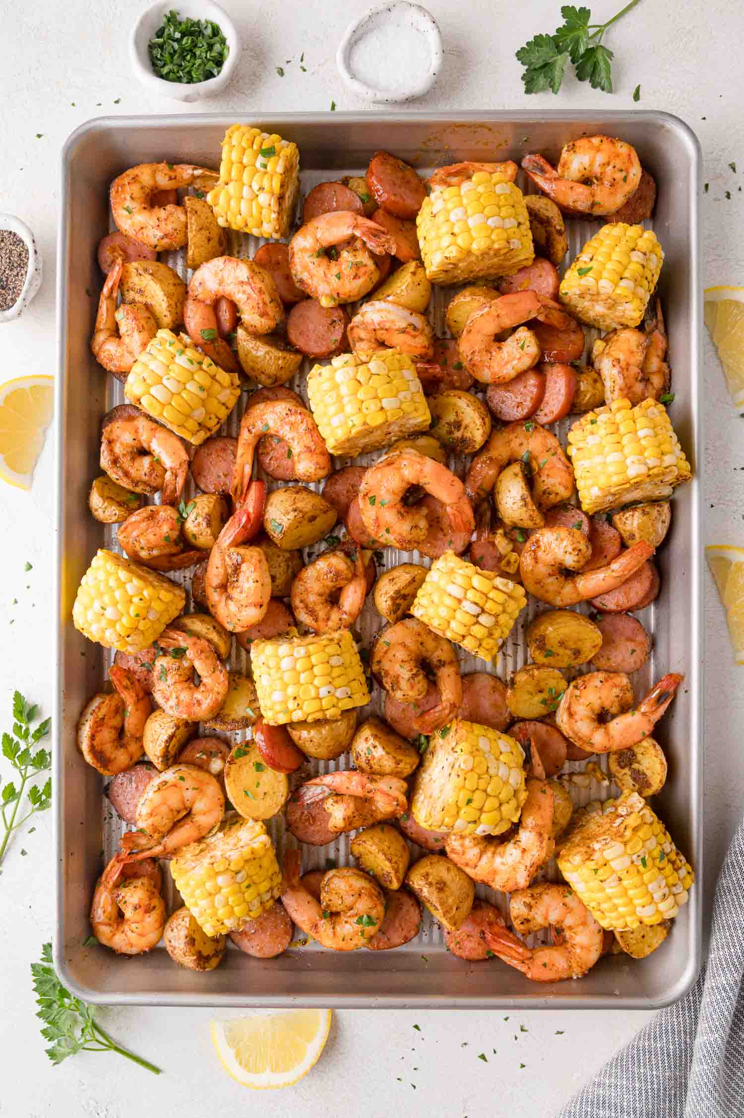 Sheet pan shrimp boil with cooked shrimp, sausage slices, corn on the cob pieces, and halved potatoes, seasoned and garnished with herbs.
