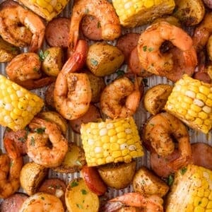 A sheet pan filled with cooked shrimp, sliced sausage, corn on the cob, and roasted potatoes, all seasoned and garnished with herbs.