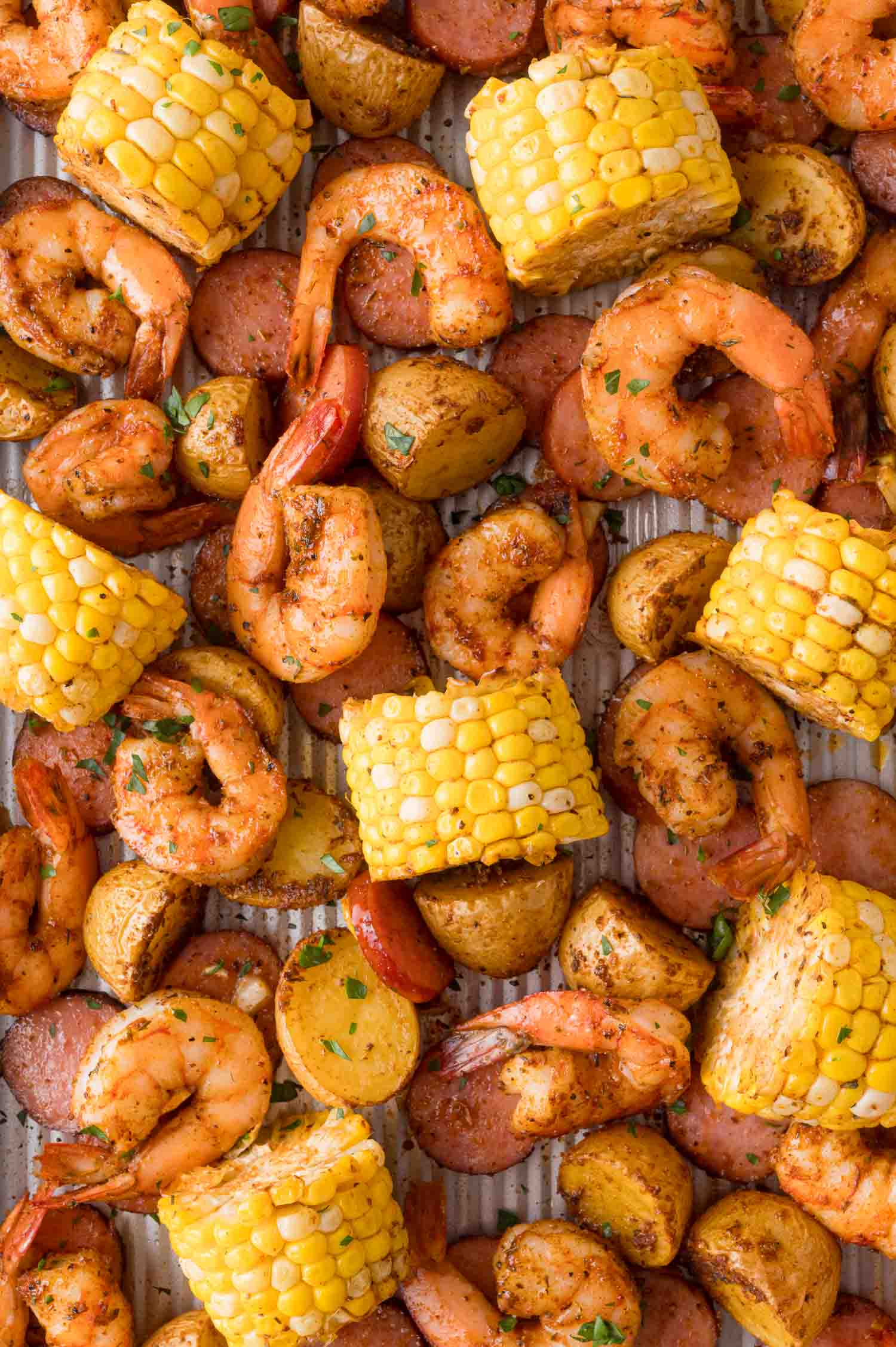 A sheet pan filled with roasted shrimp, sliced sausage, corn on the cob pieces, and halved baby potatoes, seasoned with herbs and spices.
