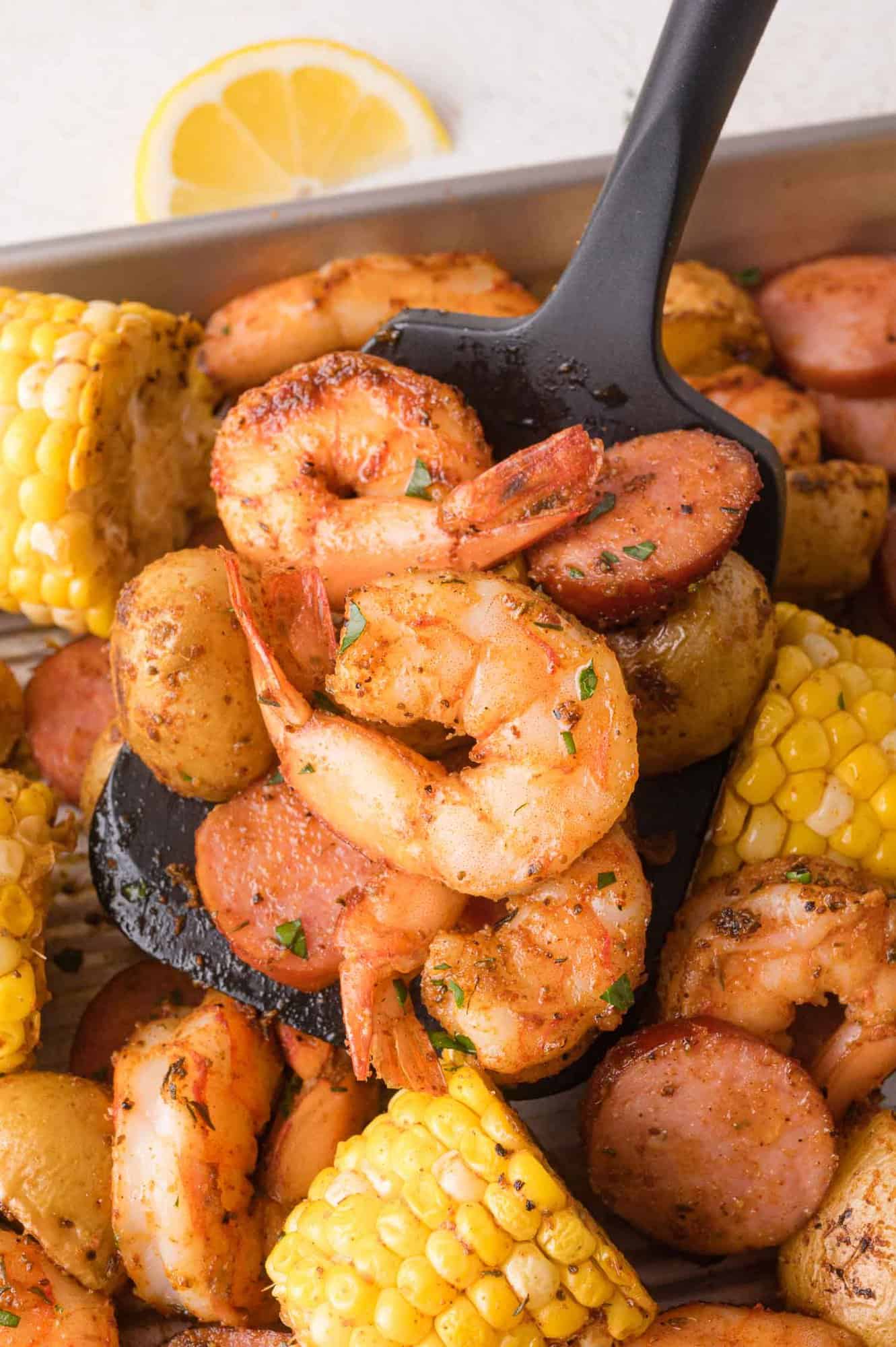 A black serving spoon holds seasoned shrimp, sliced sausage, potatoes, and corn from a tray of cooked seafood and vegetables. A lemon wedge is in the background.