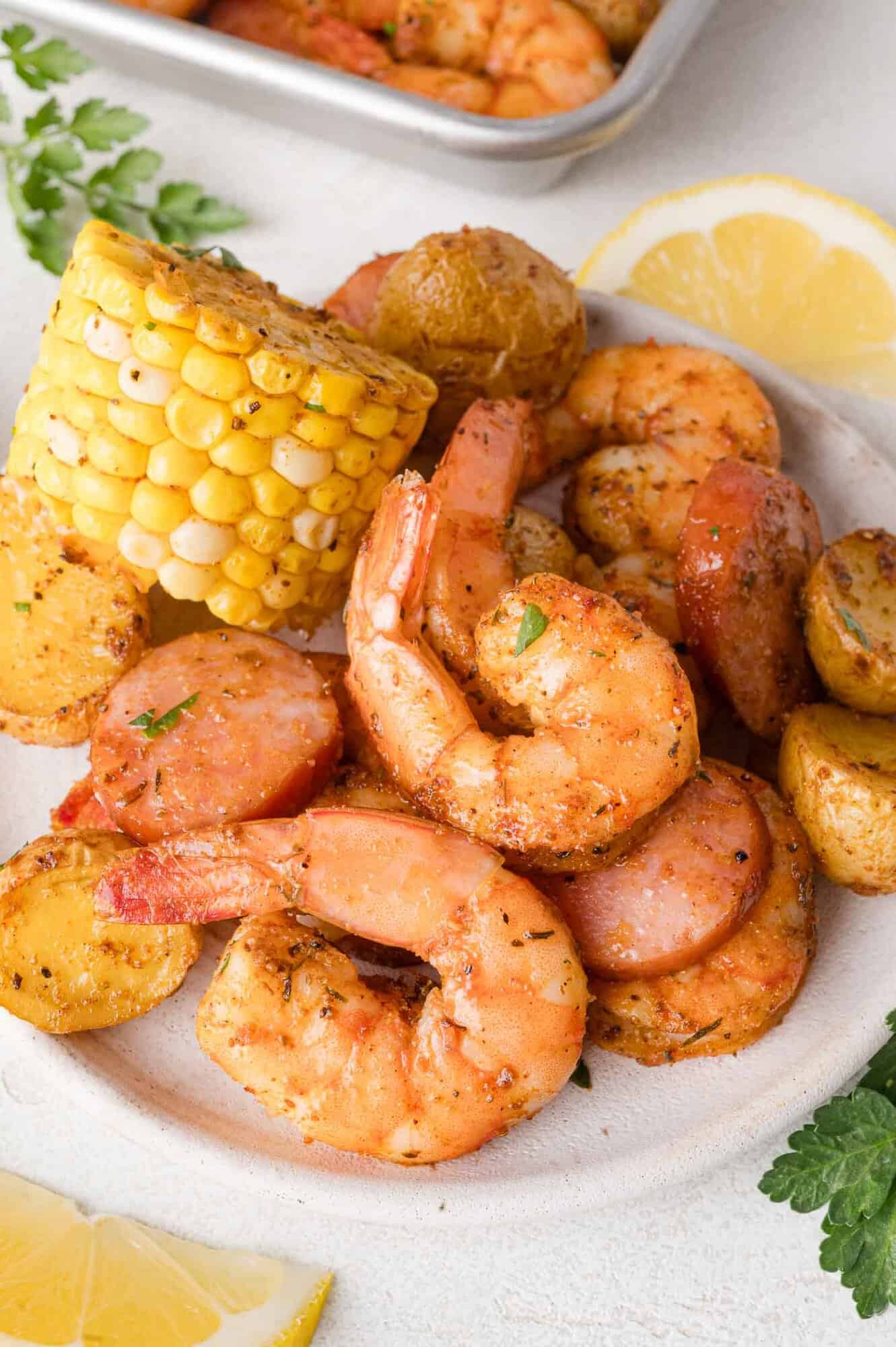 Plate of cooked shrimp, sausage slices, corn on the cob, and seasoned potatoes, garnished with herbs, with lemon slices and parsley on the side.