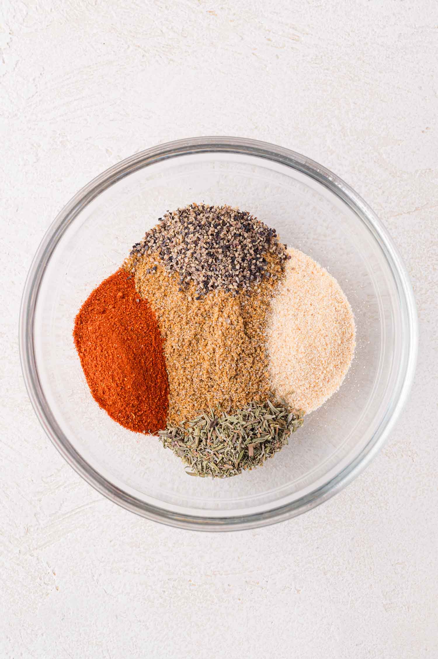 A glass bowl containing separate piles of dried spices, including paprika, black pepper, garlic powder, thyme, and brownish seasonings, on a light surface.