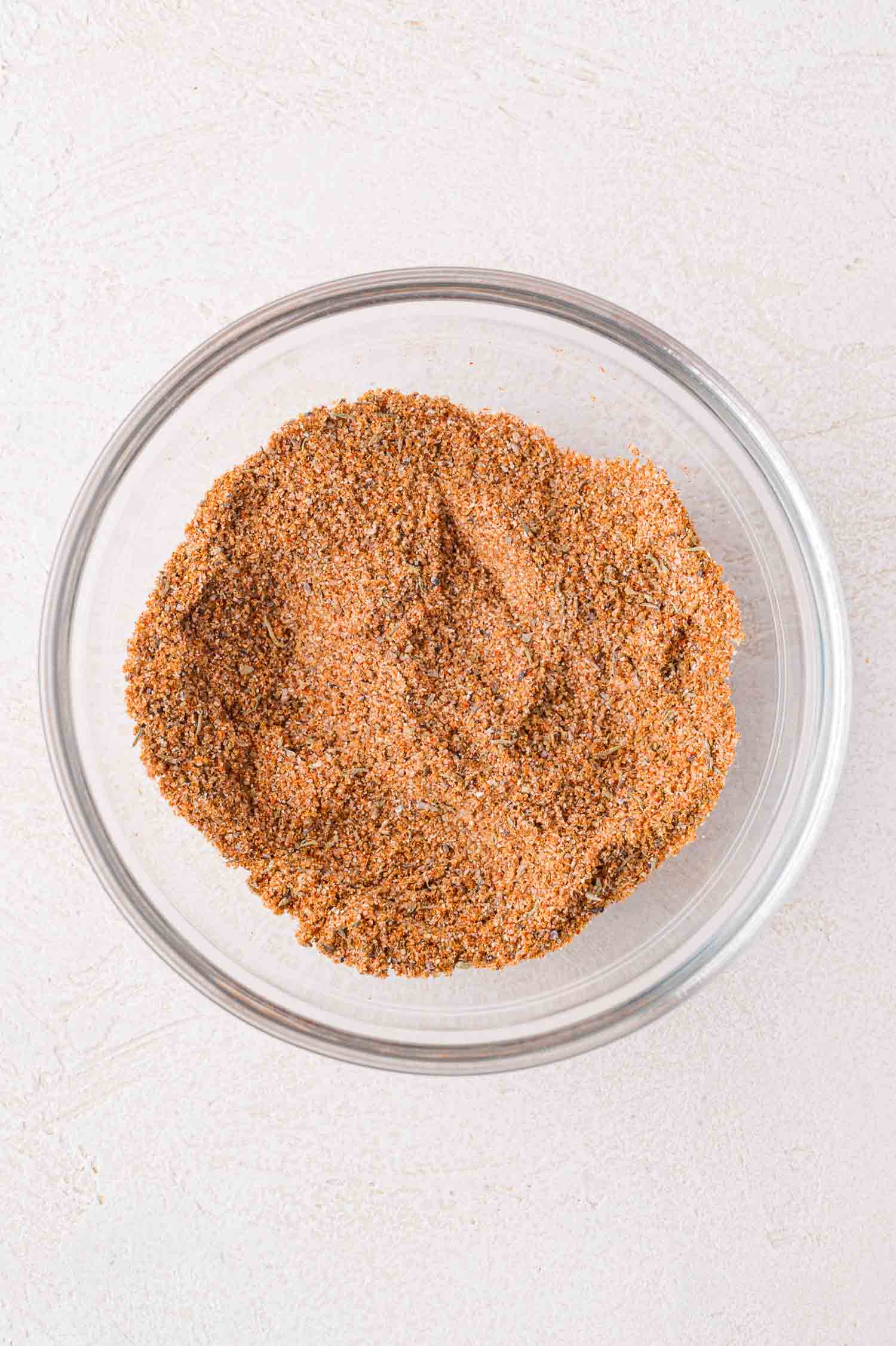 A glass bowl filled with a mixed blend of dry spices and seasonings on a light-colored surface.