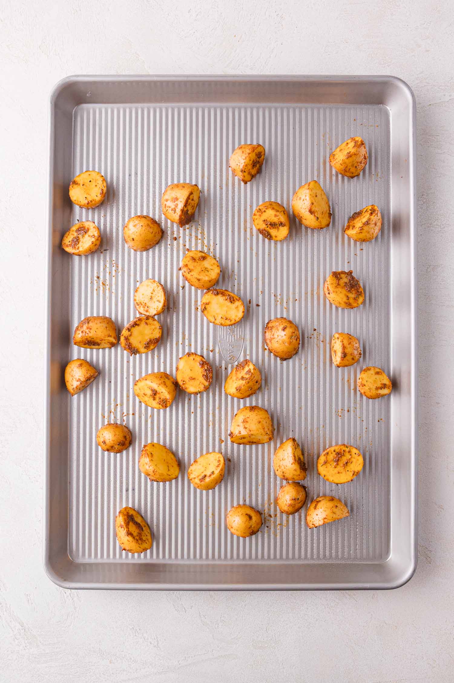 A baking sheet with evenly spaced, halved baby potatoes seasoned with spices, ready to be roasted.
