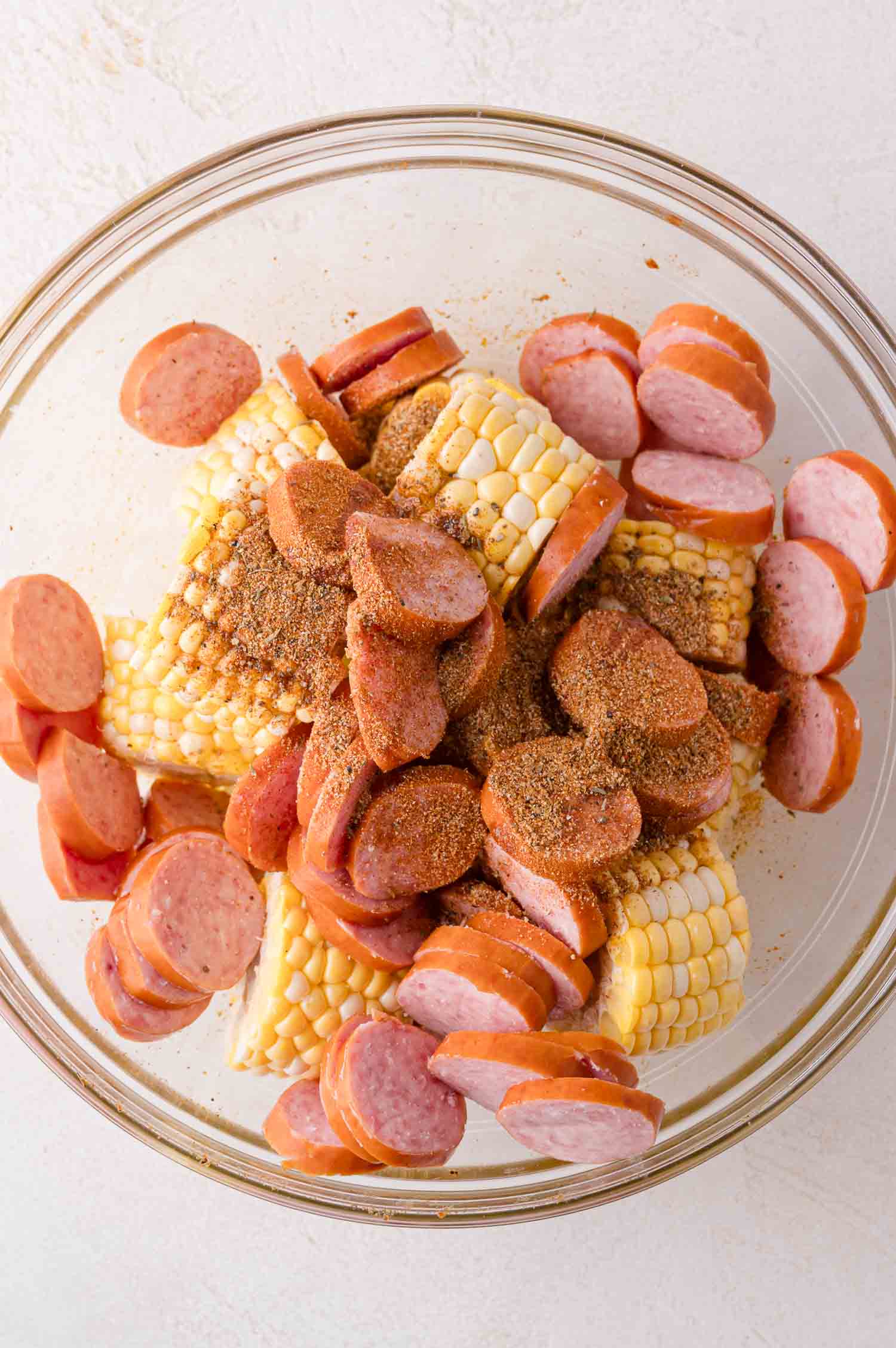 A glass bowl containing sliced sausage, corn on the cob pieces, and seasoning on a white surface.