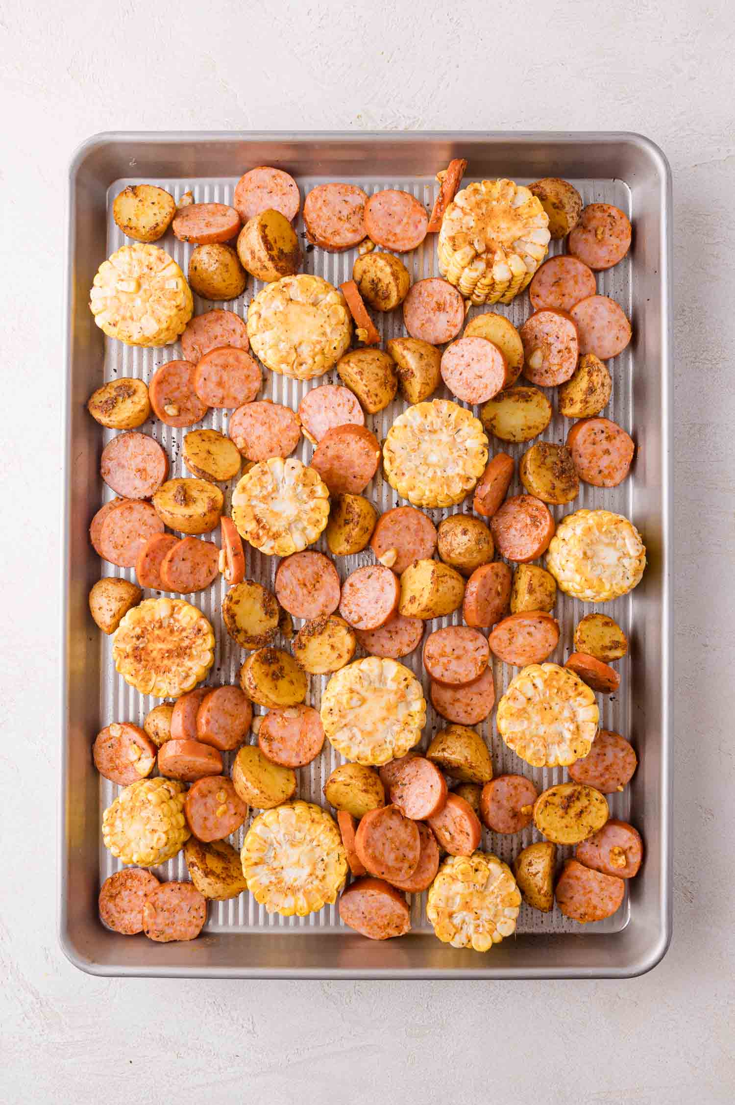 Sheet pan with sliced sausage, halved baby potatoes, and corn on the cob rounds, all seasoned and arranged evenly on a metal baking tray.