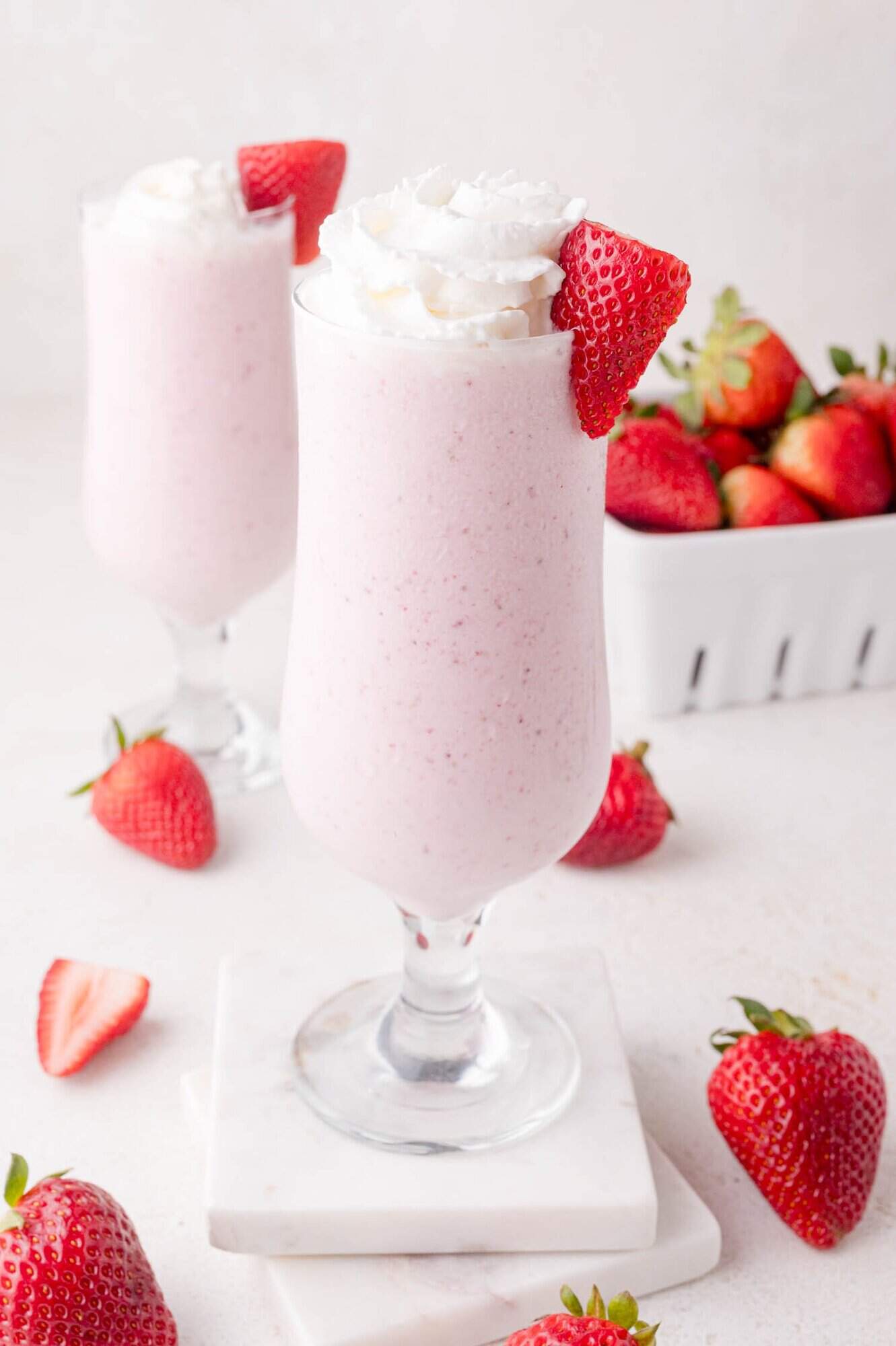 A tall glass of strawberry milkshake topped with whipped cream and a strawberry slice, with fresh strawberries scattered around and another milkshake in the background.