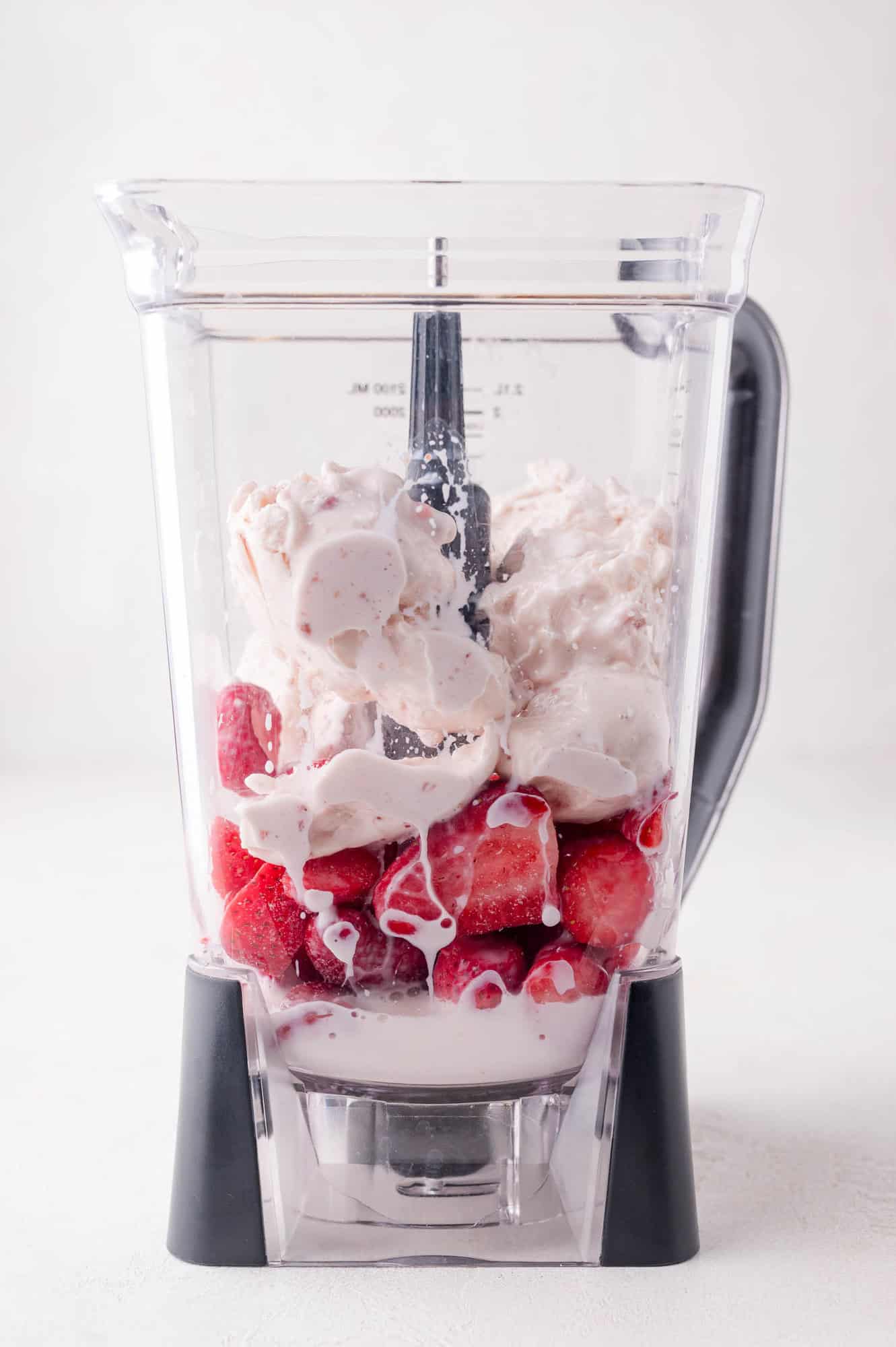 A blender filled with frozen strawberries, dollops of vanilla ice cream, and a small amount of milk, ready to be blended.