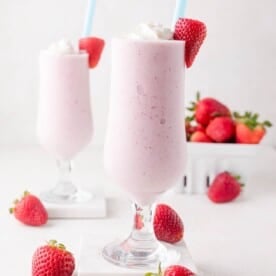 Two glasses of strawberry milkshake topped with whipped cream and a fresh strawberry, with whole strawberries scattered around and a container of strawberries in the background.