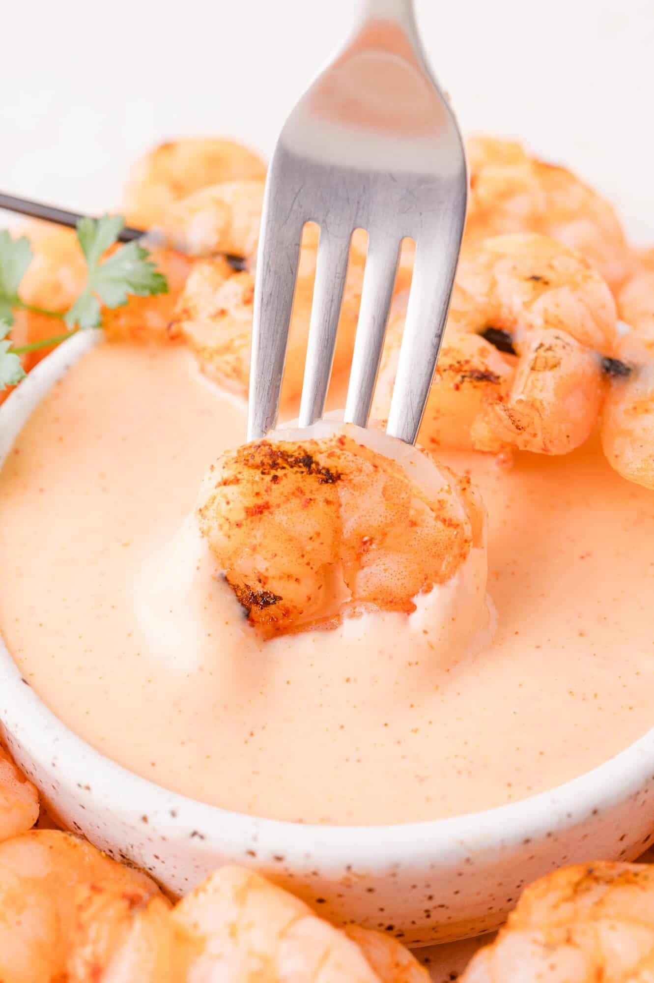 A grilled shrimp held by a fork is being dipped into a bowl of creamy orange sauce, with more shrimp and a sprig of parsley in the background.