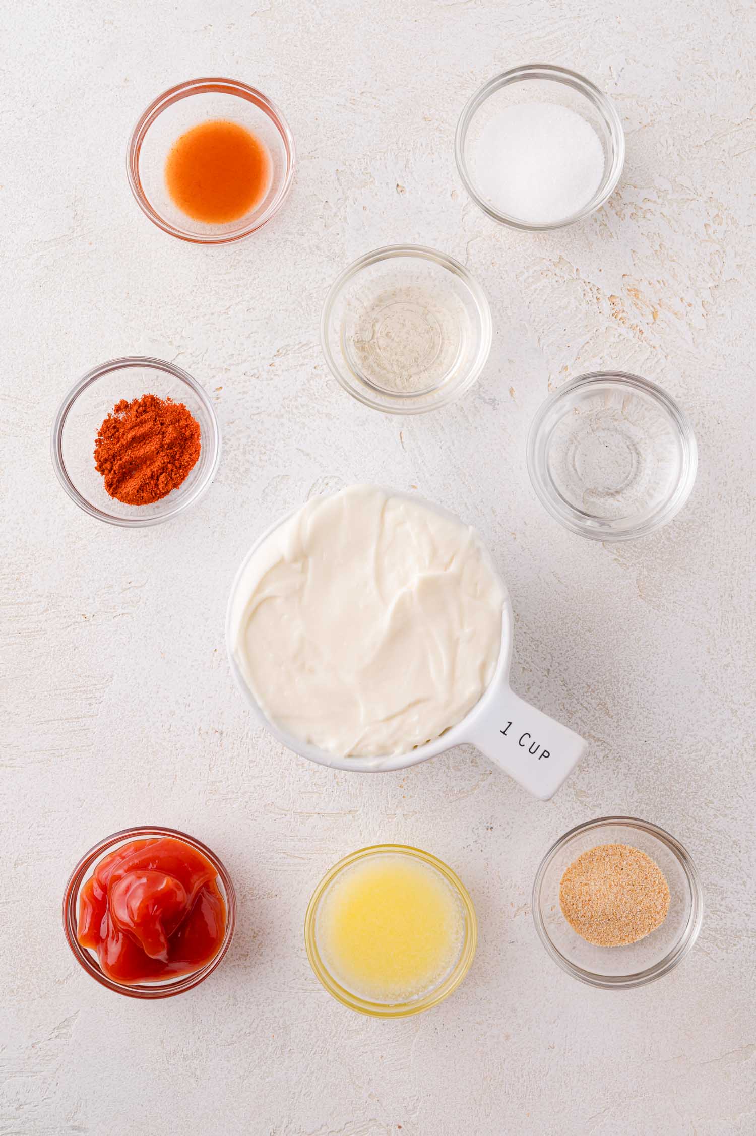 Nine small bowls containing various ingredients, including mayonnaise, ketchup, hot sauce, spices, and liquids, arranged on a white surface.