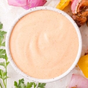 A white bowl filled with creamy, light orange yum yum sauce, surrounded by chopped parsley, onions, and yellow bell pepper pieces.