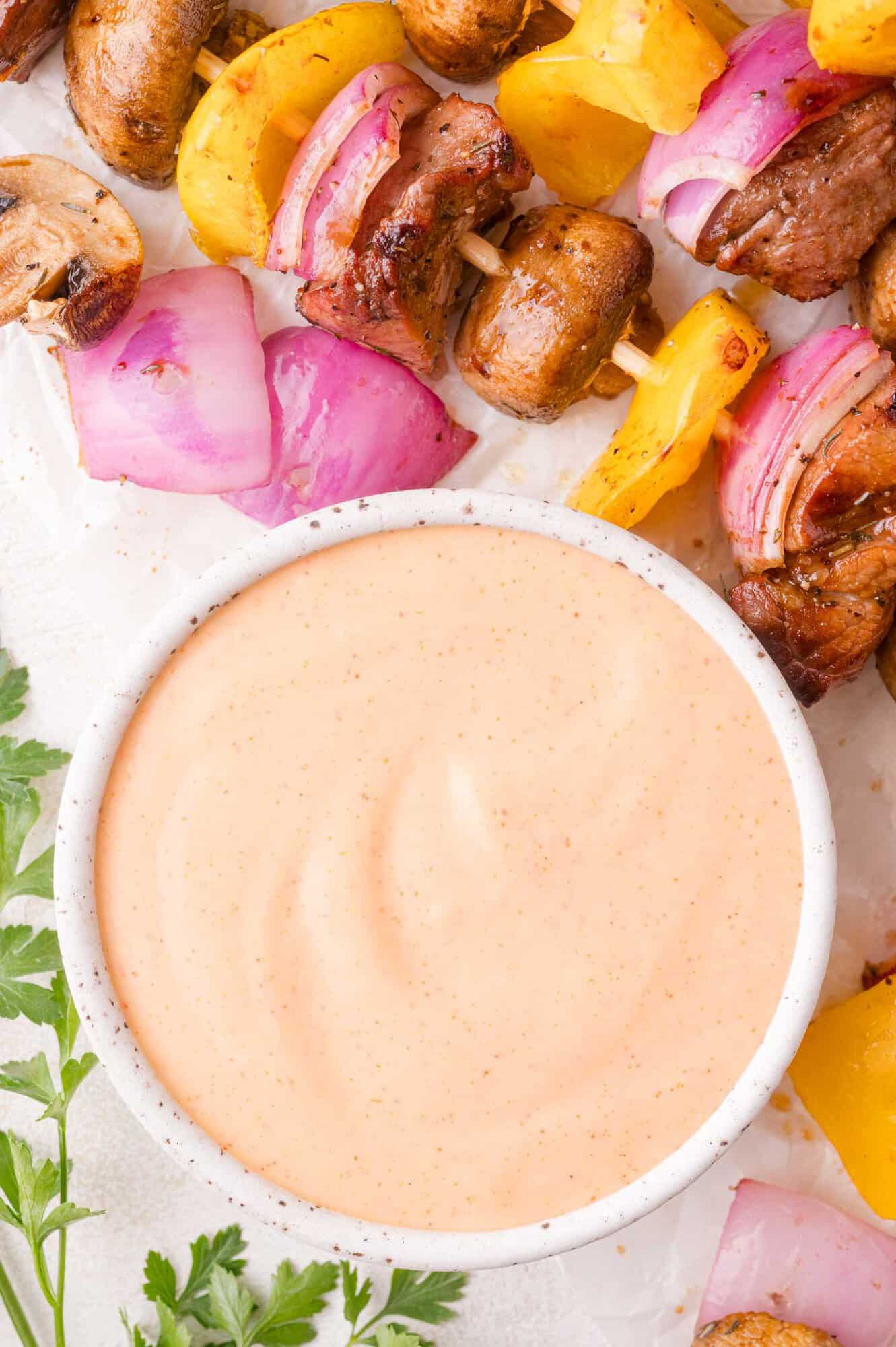 A bowl of creamy dipping yum yum sauce sits next to grilled vegetable and meat skewers with mushrooms, red onions, and yellow bell peppers. Parsley is visible in the corner.