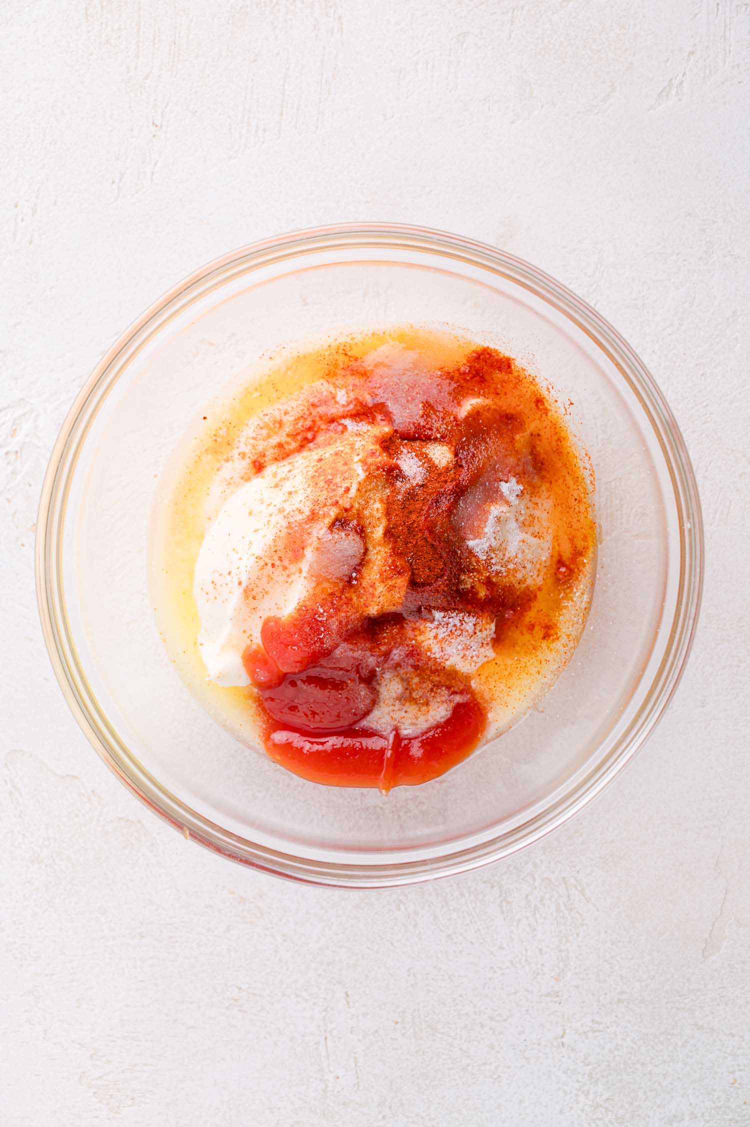 A glass bowl containing mayonnaise, ketchup, and various spices, sitting on a white surface.