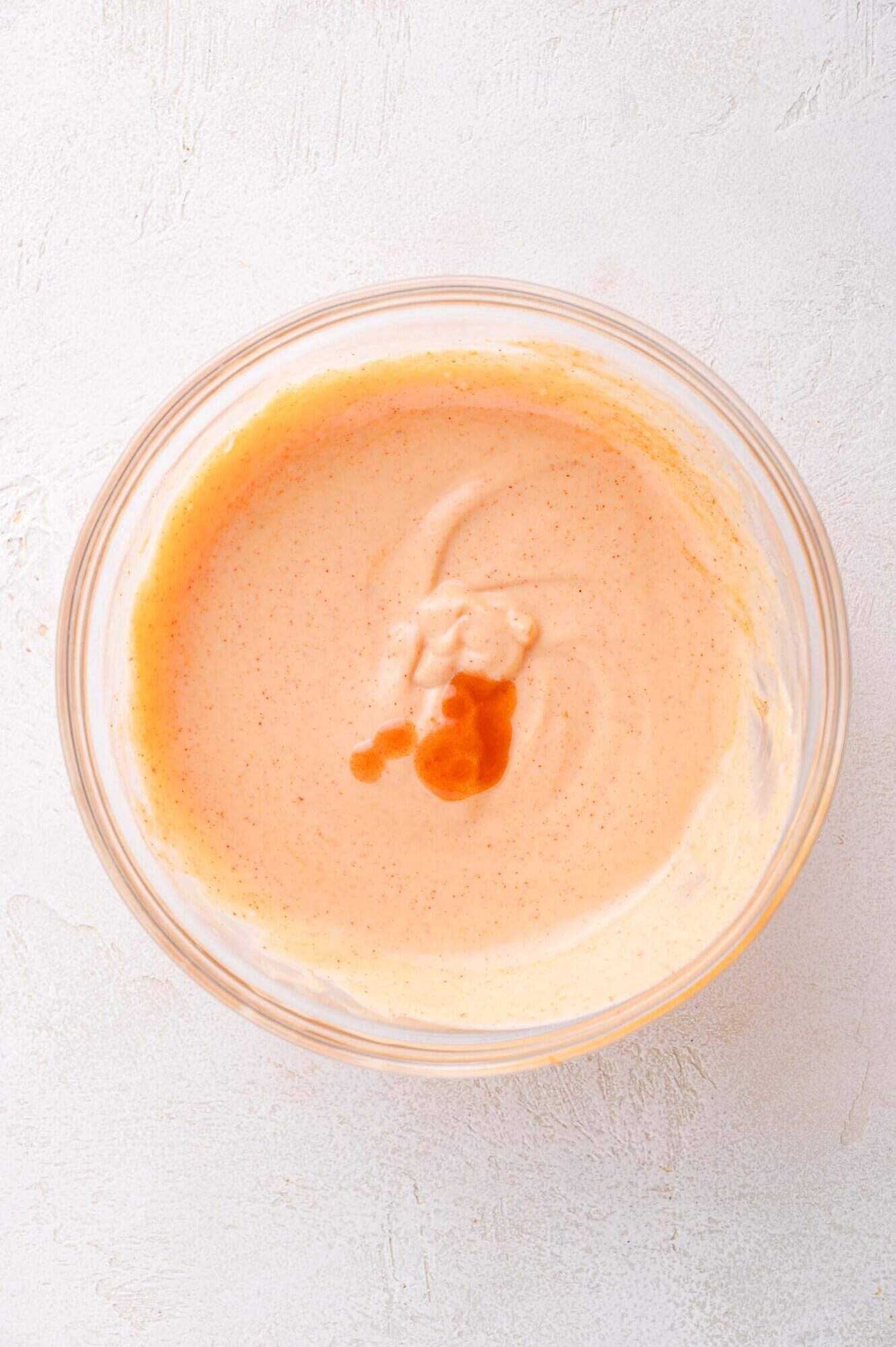 A glass bowl containing a creamy, light orange sauce with a few drops of darker liquid on top, set on a light textured surface.