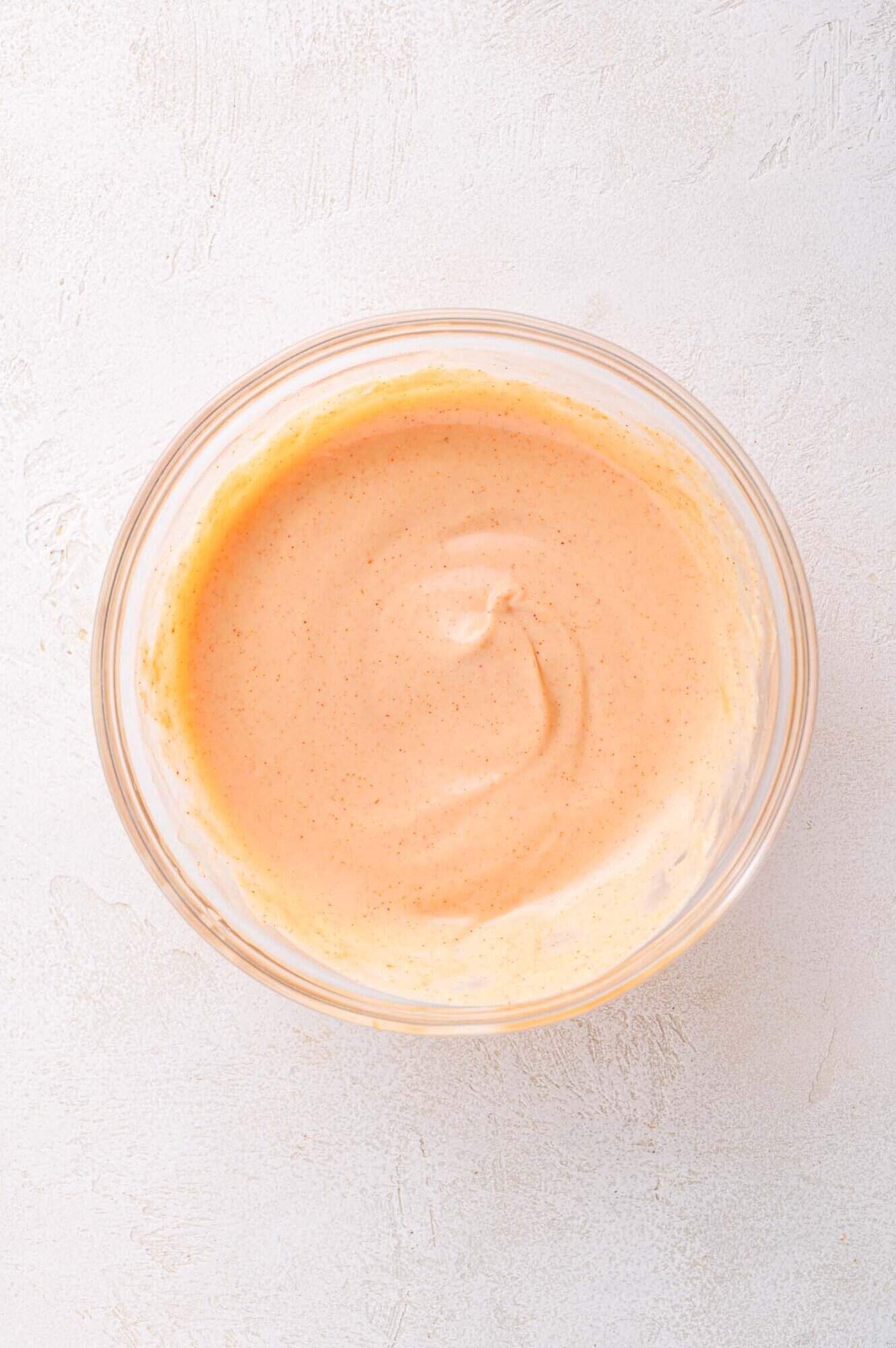 A glass bowl filled with creamy, orange-colored yum yum sauce sits on a light textured surface.