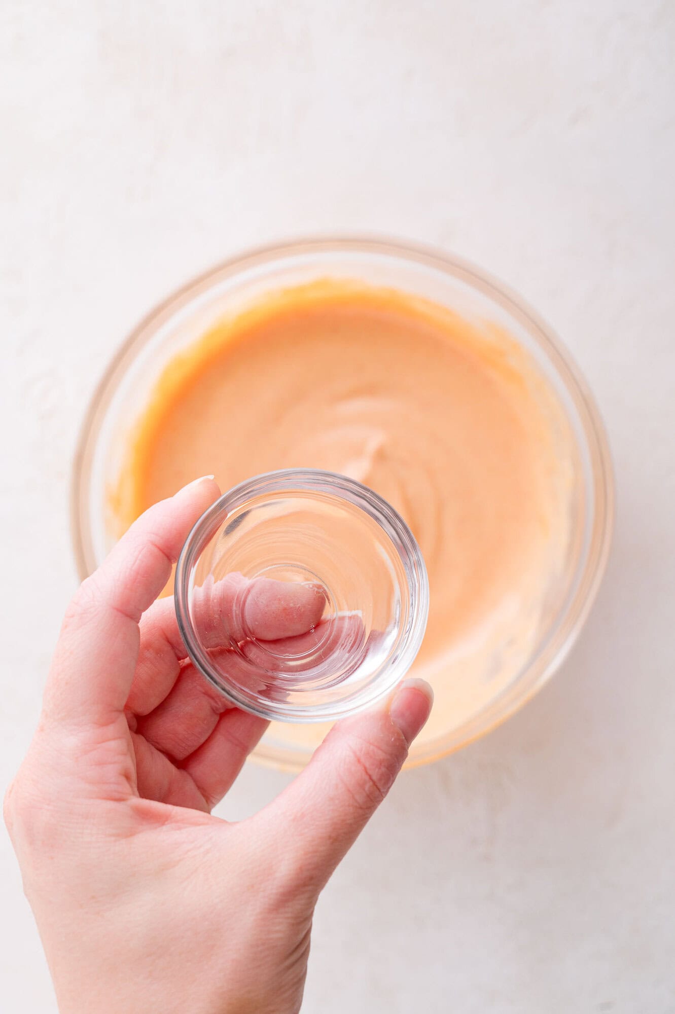 A hand holding a small clear glass bowl above a larger glass bowl containing an orange mixture on a light surface.
