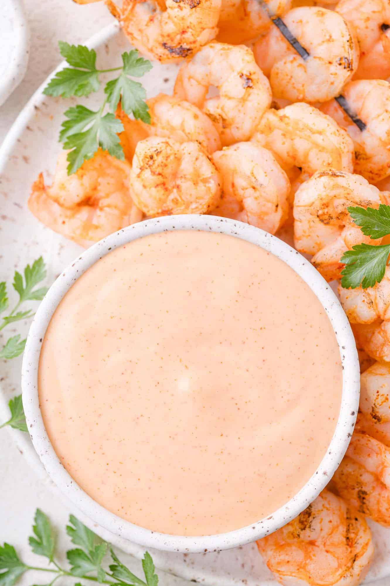 A bowl of creamy orange dipping sauce is surrounded by grilled shrimp and fresh parsley on a white plate.