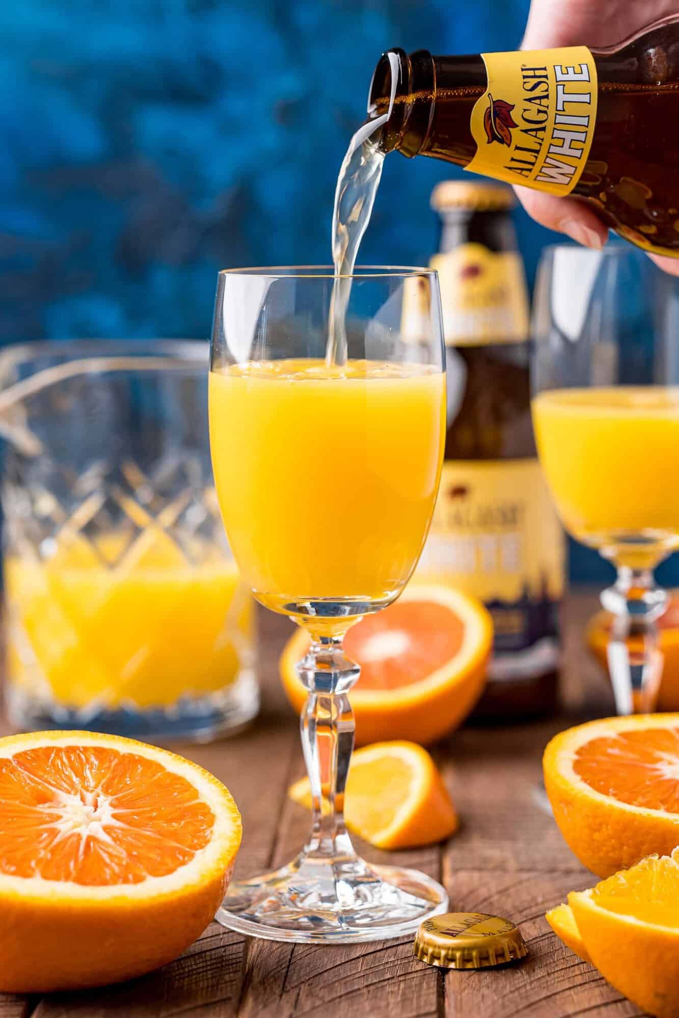 A hand pours Allagash White beer into a glass of orange juice on a wooden table, surrounded by sliced oranges, another glass, and a beer bottle.