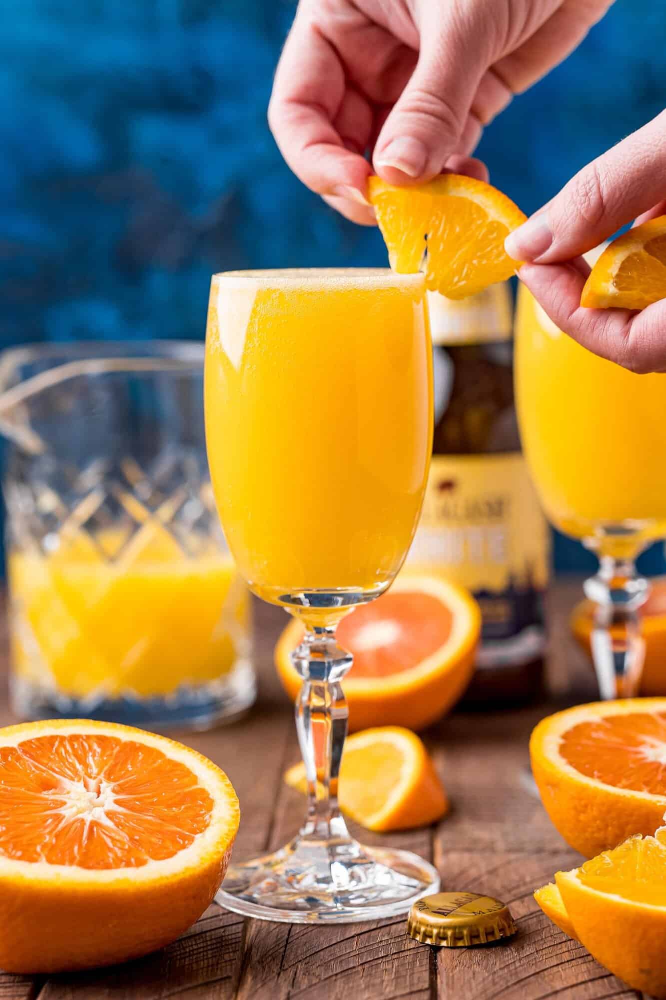 A hand garnishes a tall glass of orange beermosa with an orange slice; fresh oranges and a bottle are on a wooden table.