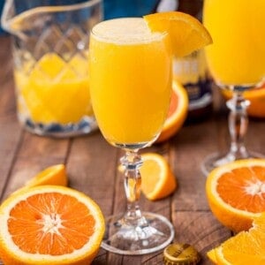 A glass of orange beermosa garnished with an orange slice sits on a wooden table with sliced oranges and a pitcher in the background.