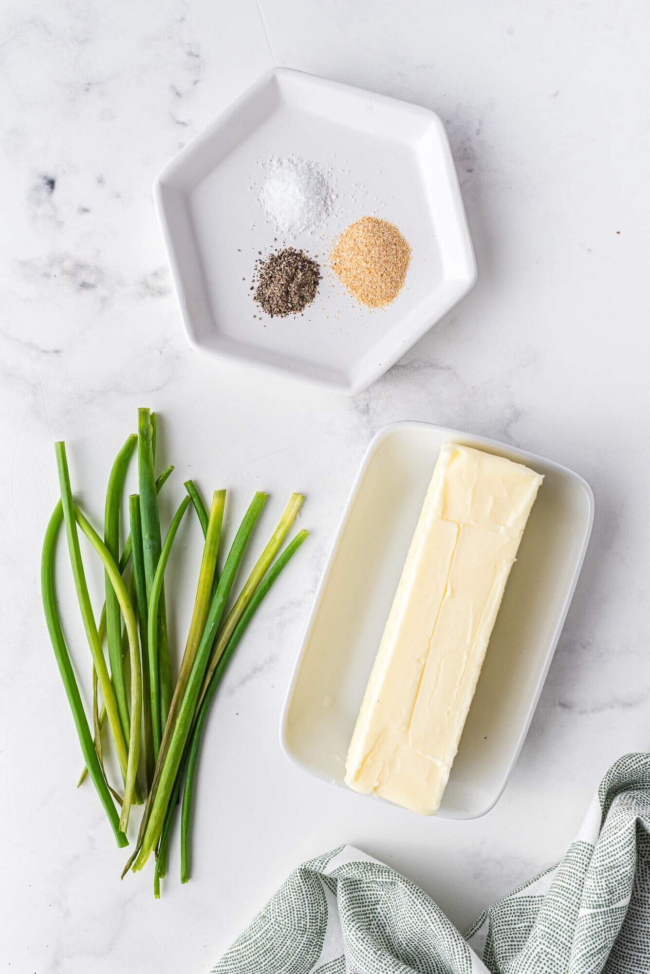 A stick of butter on a dish, chives, and a plate with salt, pepper, and garlic powder on a white marble surface.