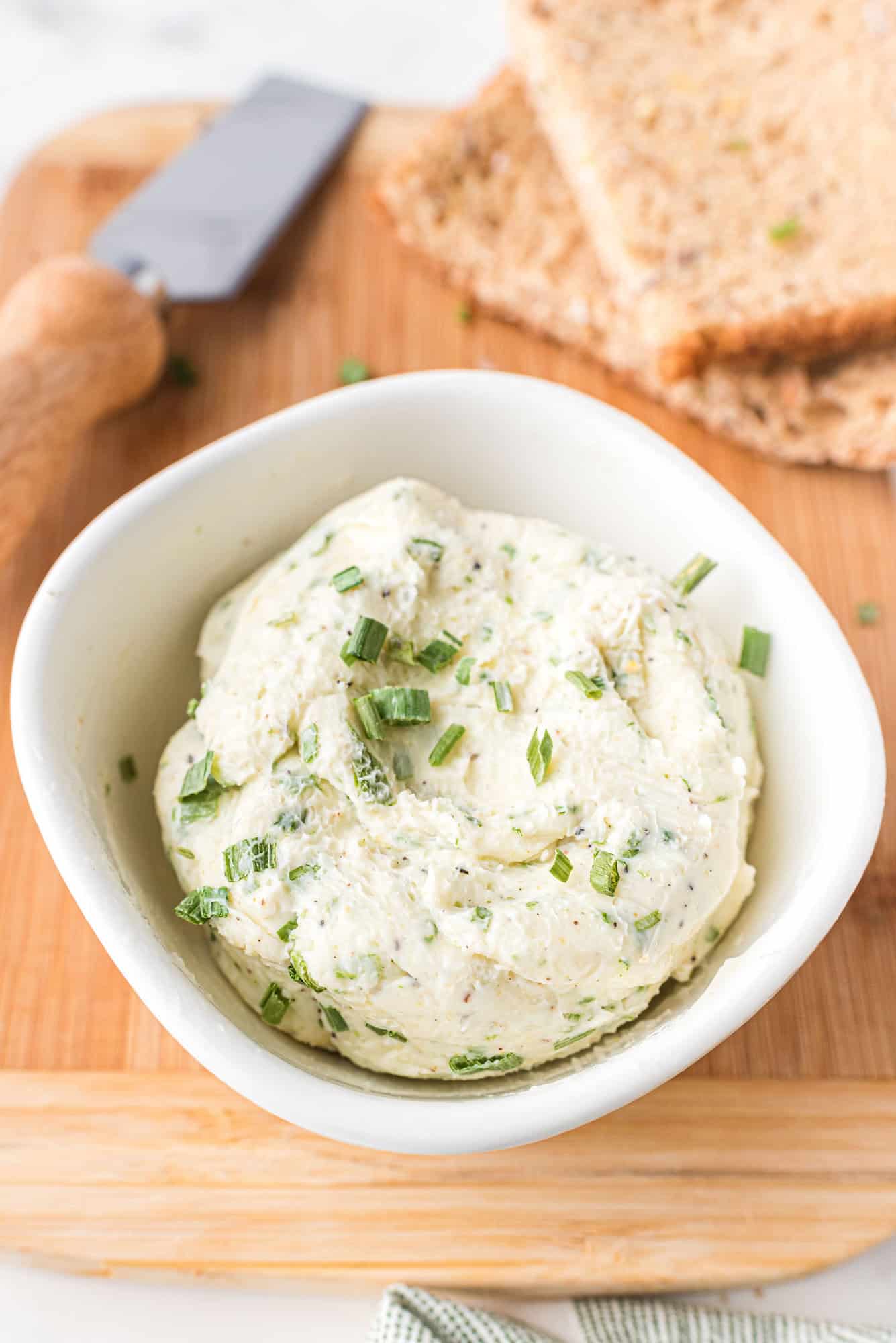 A white bowl filled with chive butter, garnished with chopped chives, sits on a wooden board next to slices of whole grain bread and a butter knife.