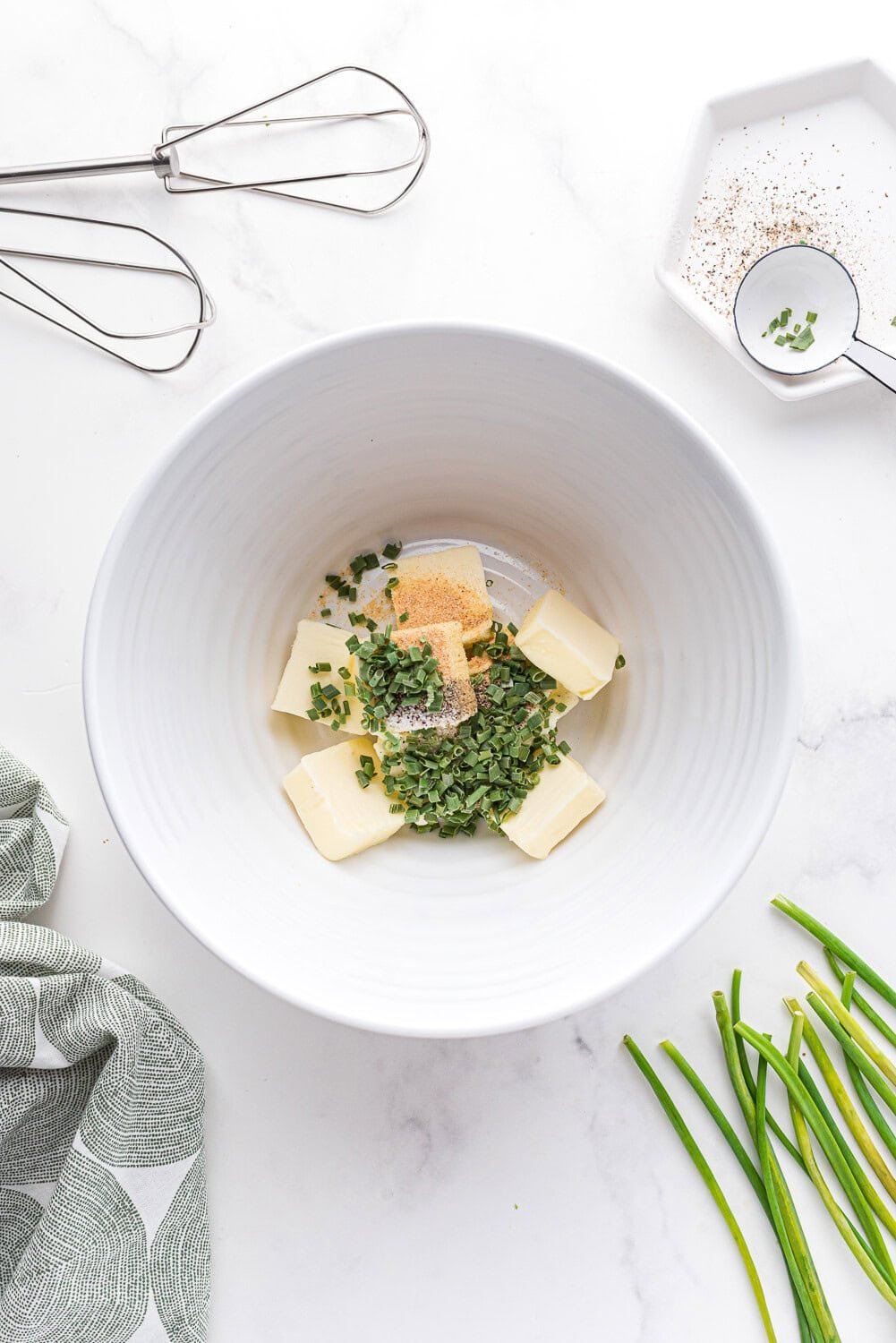 Large white bowl with chunks of butter, chopped chives, garlic powder, salt, and pepper. Whisks, a towel, and fresh chives are nearby on a white countertop.