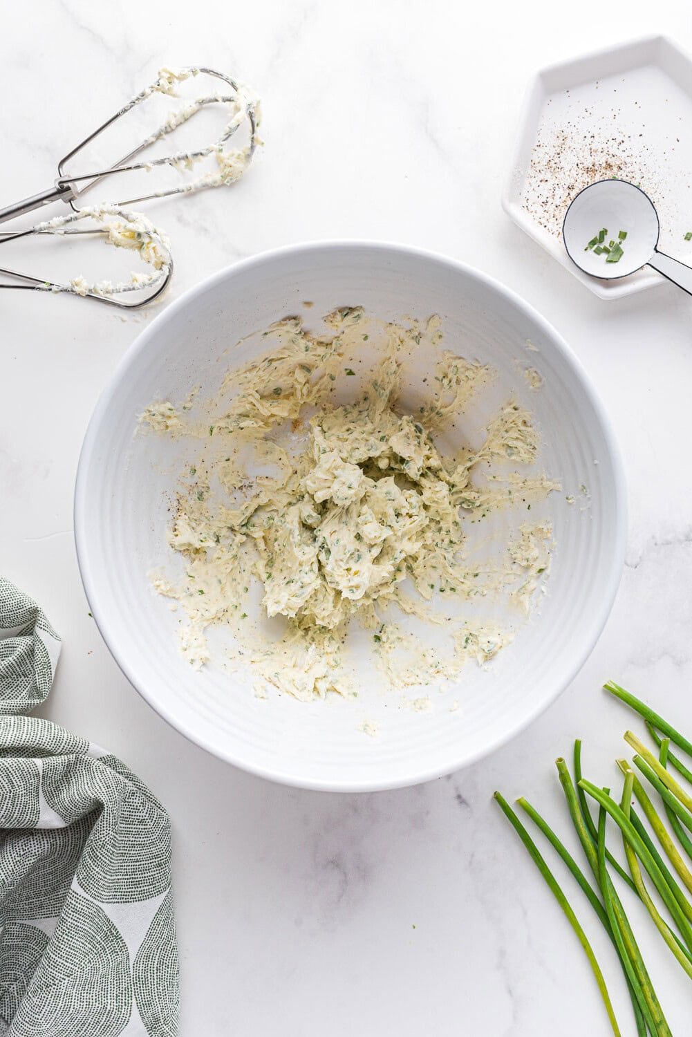 A white bowl with a mixture of whipped chive butter sits on a marble surface, surrounded by a hand mixer, a small plate of spices, chives, and a patterned cloth.