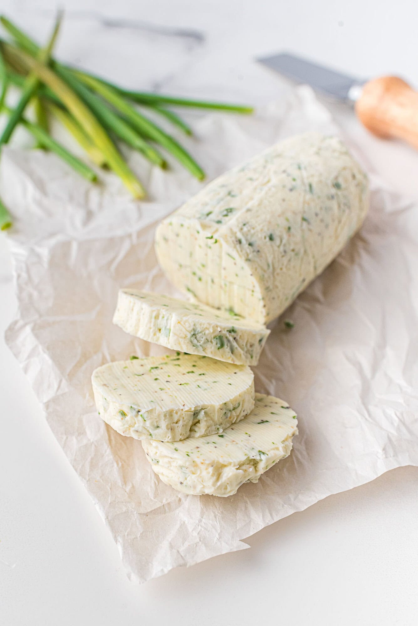 A log of compound butter with chives is partially sliced on parchment paper, with a knife and fresh chives in the background.