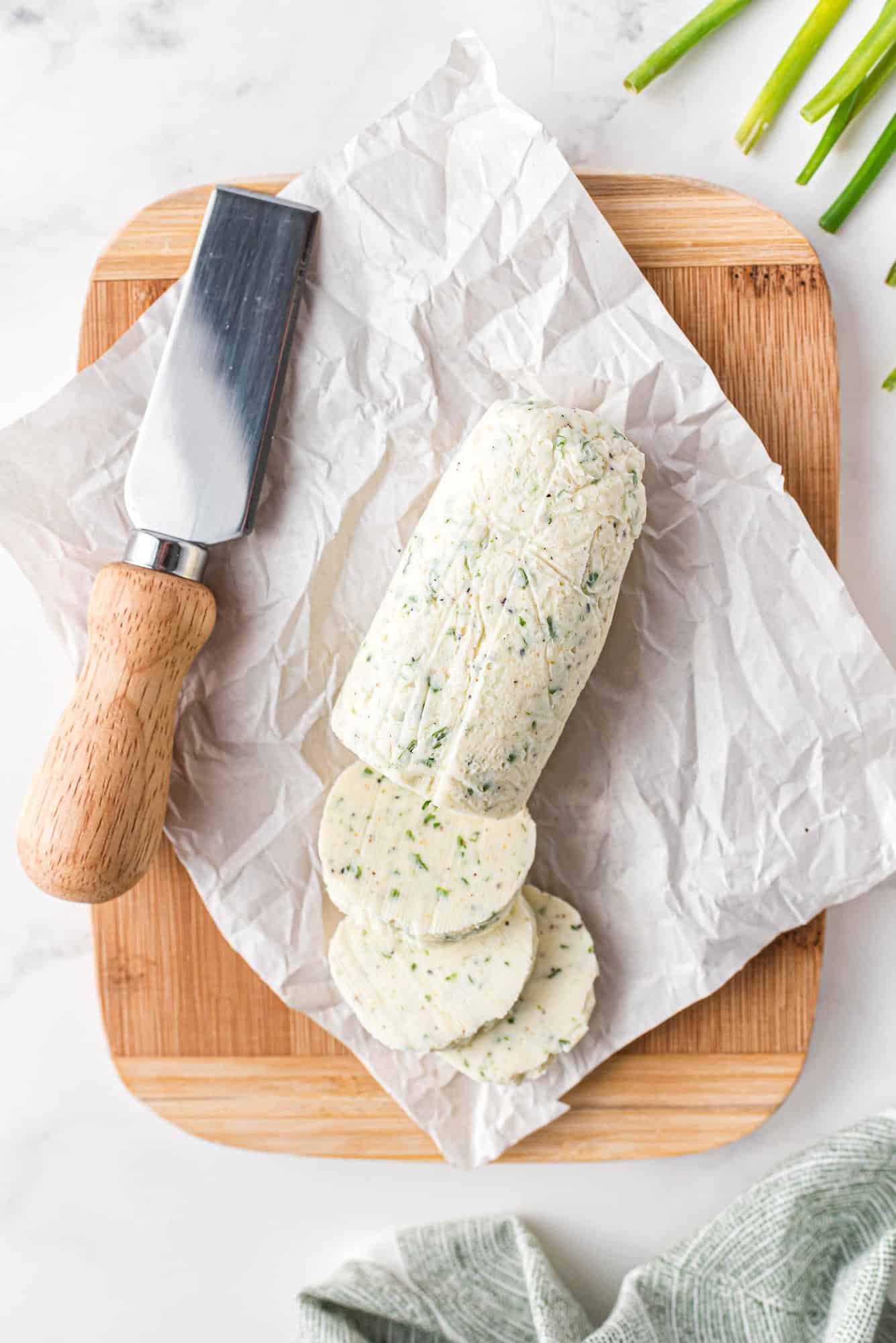 A partially sliced log of herb butter on parchment paper sits on a wooden cutting board next to a butter knife with a wooden handle.