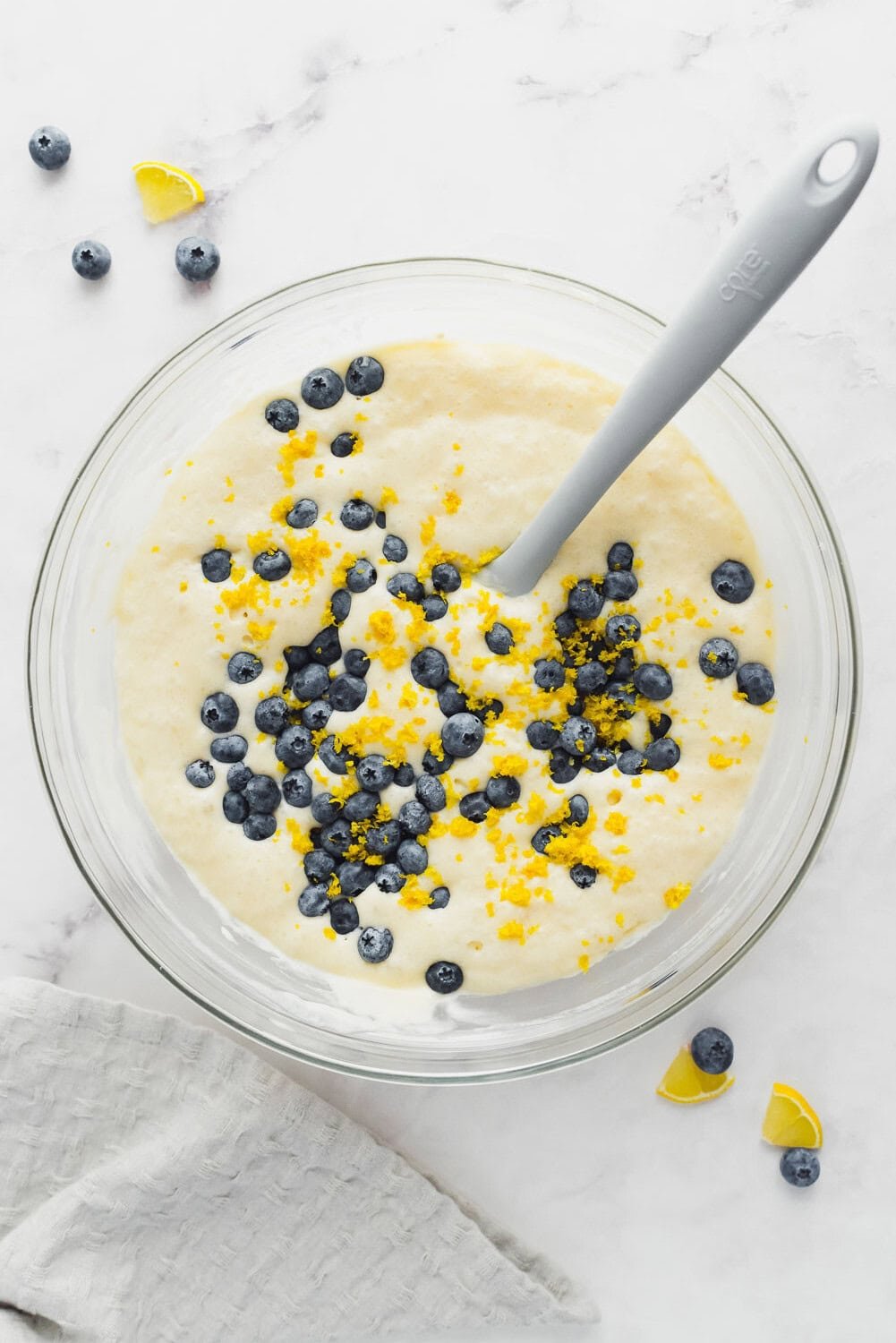 A glass bowl filled with pancake batter, topped with fresh blueberries and lemon zest, with a gray spoon resting inside. Blueberries and lemon wedges are scattered nearby on a white surface.
