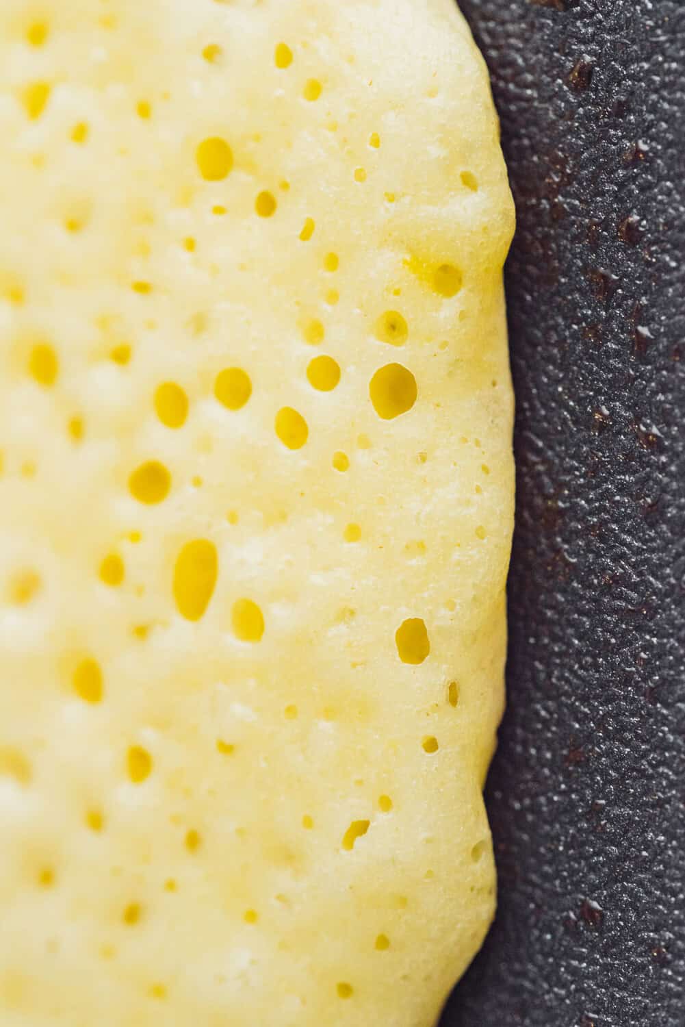 Close-up of a yellow pancake with visible bubbles cooking on a dark, textured surface.