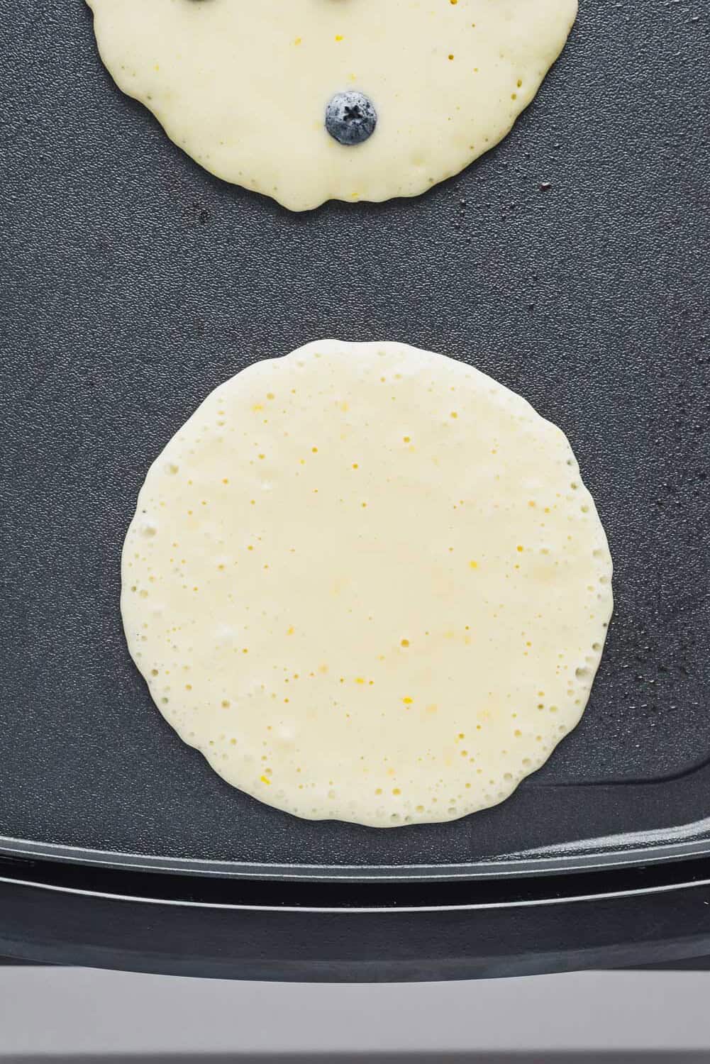 Two pancake batters cooking on a griddle, one with a blueberry visible and the other plain.