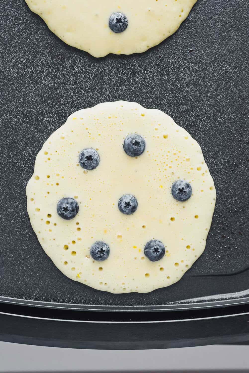 A round pancake with blueberries embedded in the batter cooks on a flat, nonstick griddle.