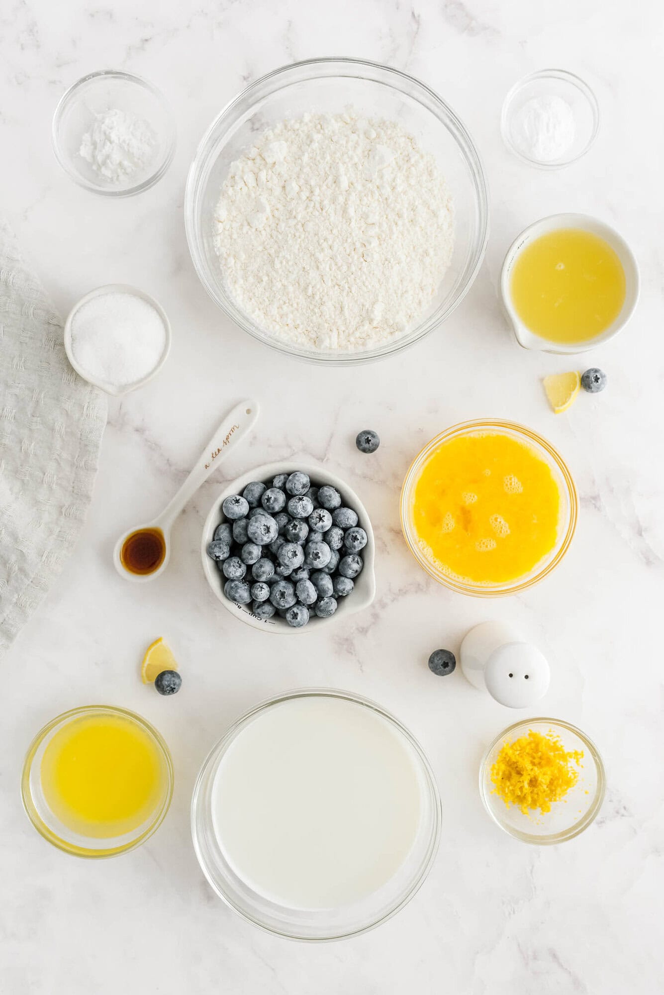 Overhead view of baking ingredients in bowls, including flour, sugar, blueberries, melted butter, milk, eggs, vanilla extract, lemon zest, and baking powder, arranged on a white surface.