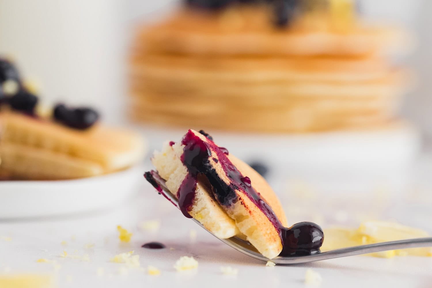 A close-up of a fork holding a piece of pancake topped with blueberry sauce, with a stack of pancakes and more sauce in the blurred background.