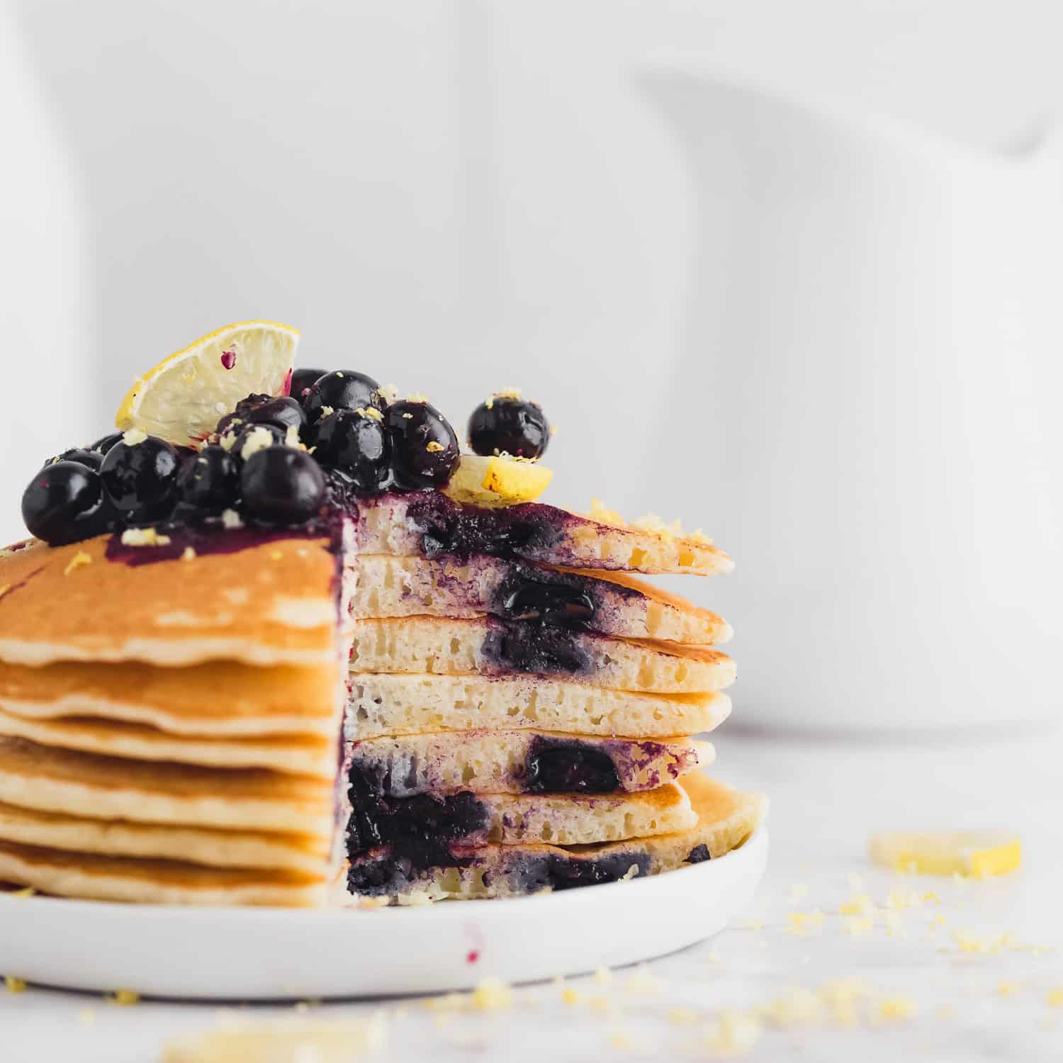 A stack of pancakes with a portion cut out, topped with blueberries, lemon slices, and lemon zest, on a white plate.