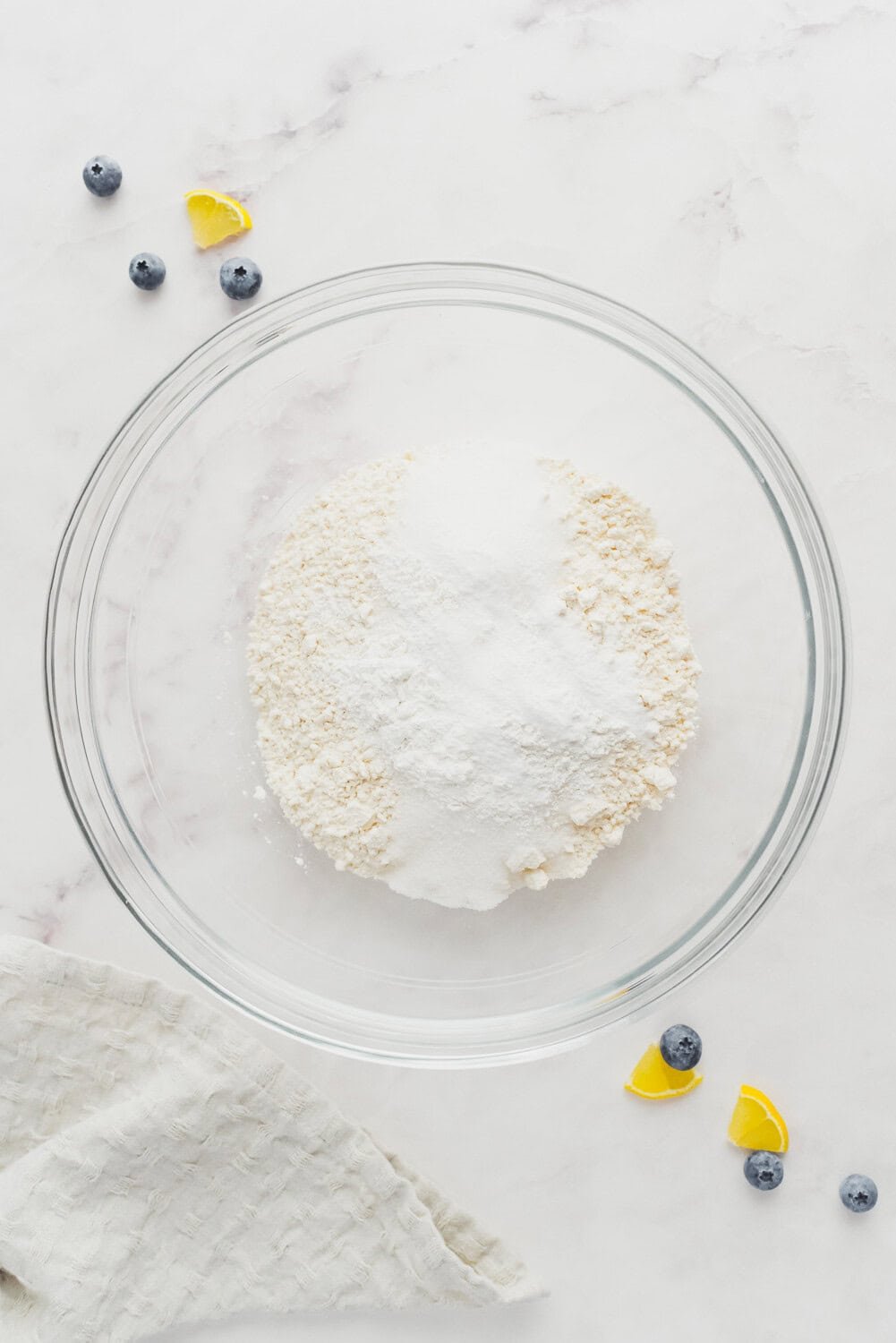 A glass bowl with flour, sugar, and baking powder on a white surface, surrounded by blueberries, lemon wedges, and a light cloth.