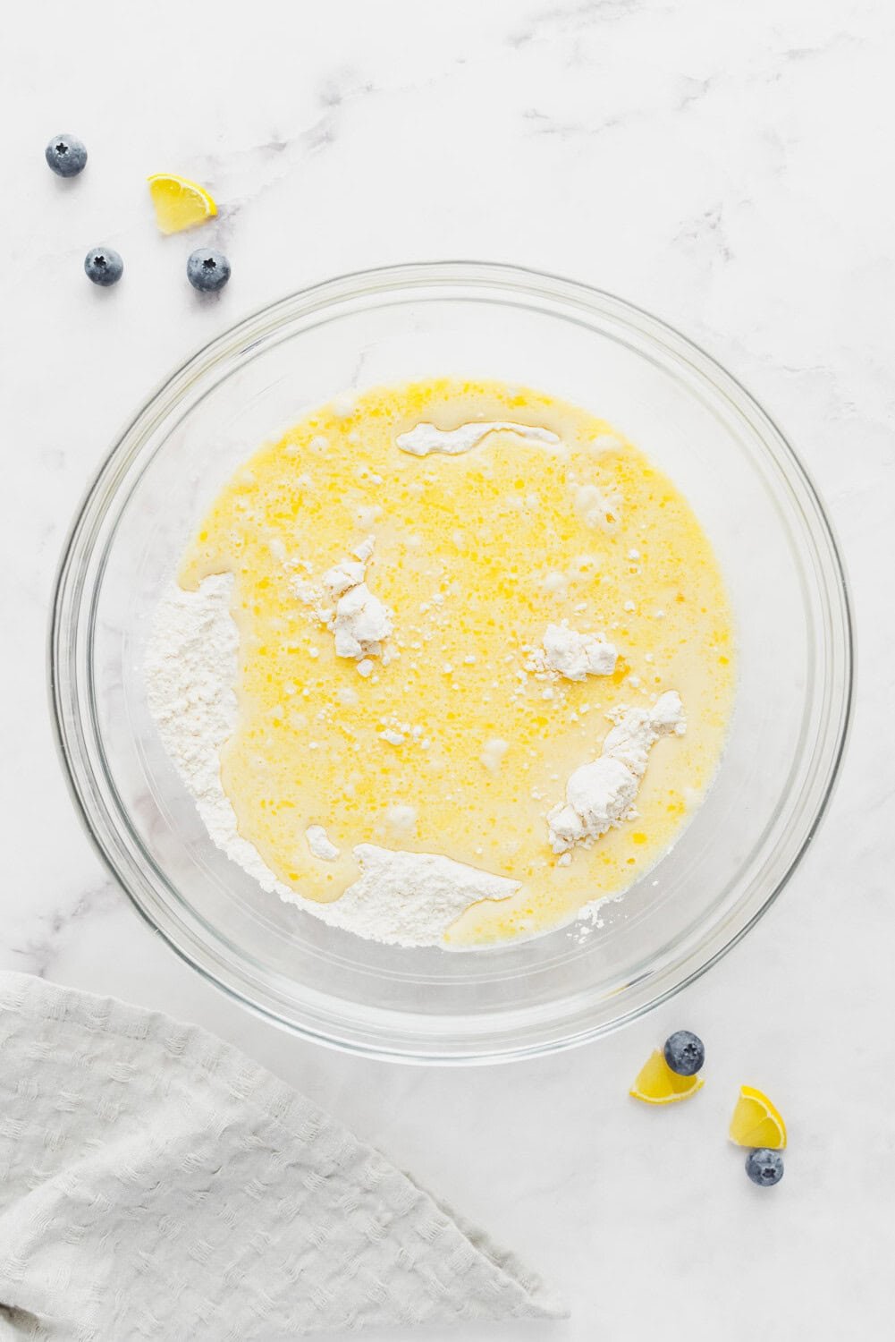 A glass bowl with flour, baking powder, wet ingredients, and eggs on a white surface, next to blueberries and lemon wedges.