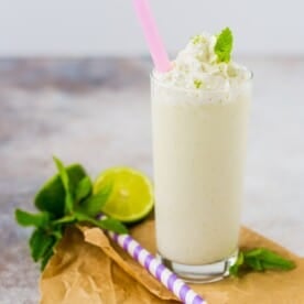 A tall glass of mojito milkshake topped with whipped cream and a mint leaf, with a pink straw. Beside it are a lime half, mint sprigs, and a purple-striped straw.